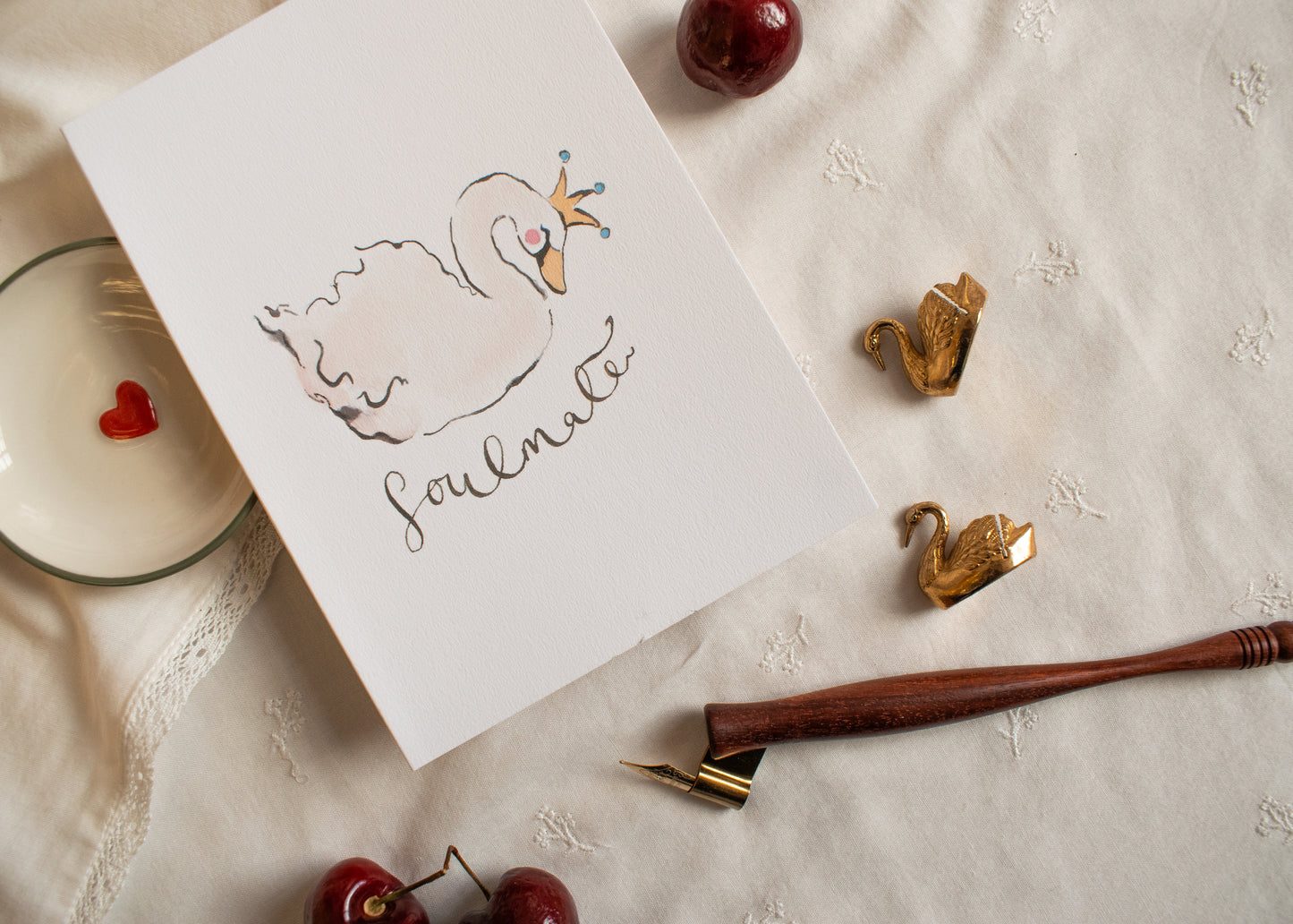 Card with a swan illustration and 'Soulmate' text, surrounded by cherries and a pen on a textured surface.