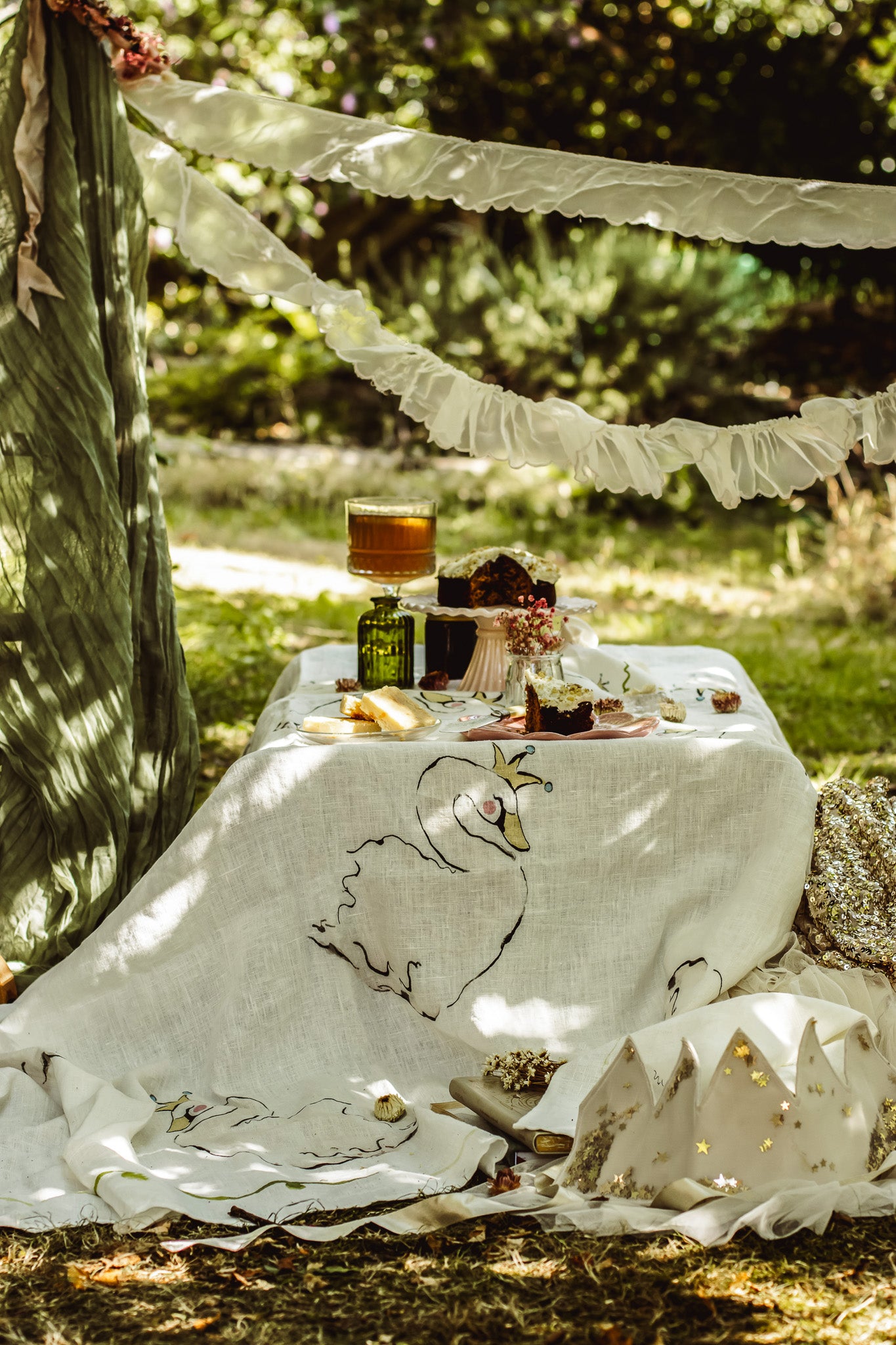 A rustic outdoor picnic setup under a tree, featuring a white linen tablecloth with embroidered swan designs, a tiered cake stand with a decorated cake, and a green bottle, set against a backdrop of green foliage and hanging white ruffled garlands.