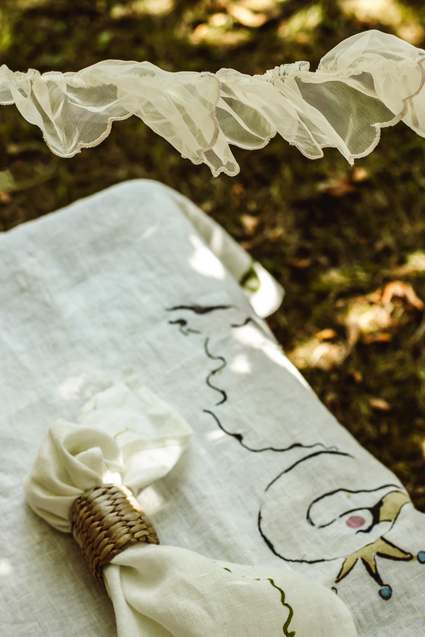 A close-up of a white embroidered fabric napkin with a wavy line drawing, tied with a woven napkin ring, resting on a matching embroidered white cloth outdoors on a grassy surface.