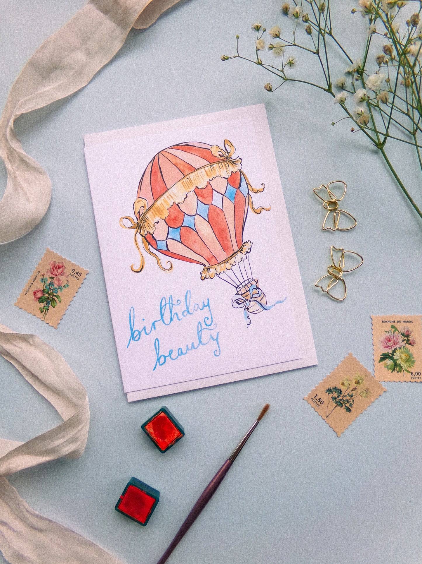 Greeting card with hot air balloon illustration and 'birthday beauty' text, surrounded by decorative items on a light blue background.