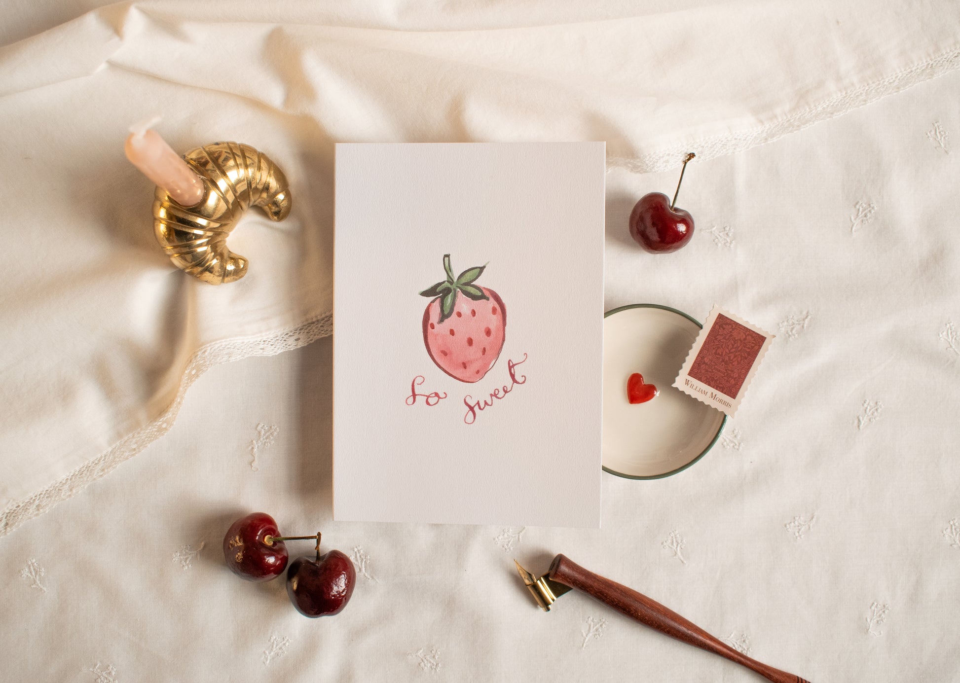 Card with strawberry illustration and 'so sweet' text on a textured surface with cherries and a pen.