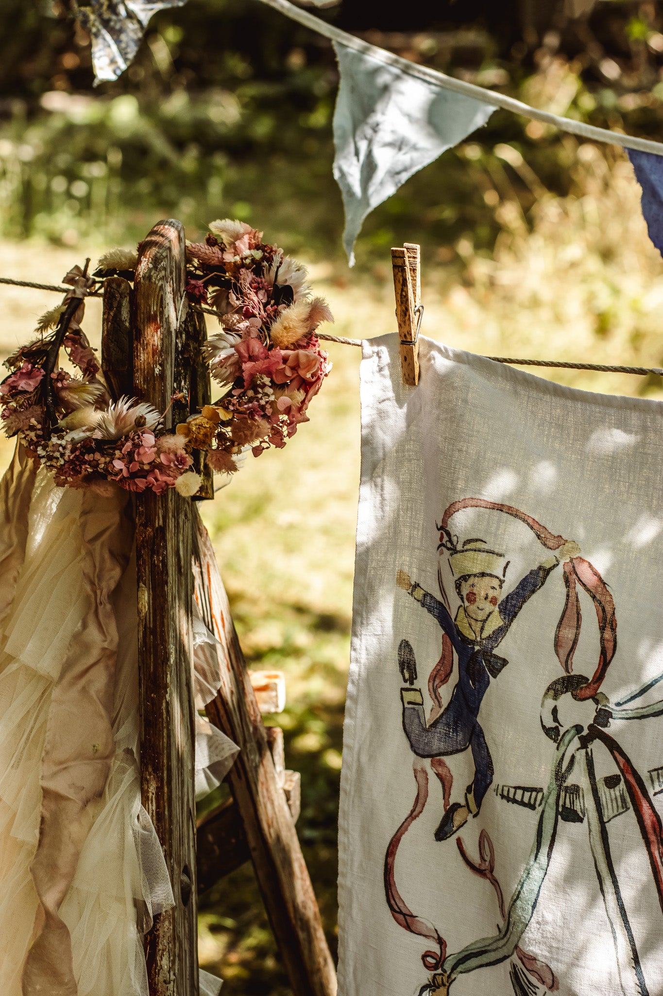 A rustic outdoor scene showing a white linen cloth with a whimsical illustration of a boy in a blue suit and hat, hanging from a wooden post with a dried floral wreath and bunting nearby.
