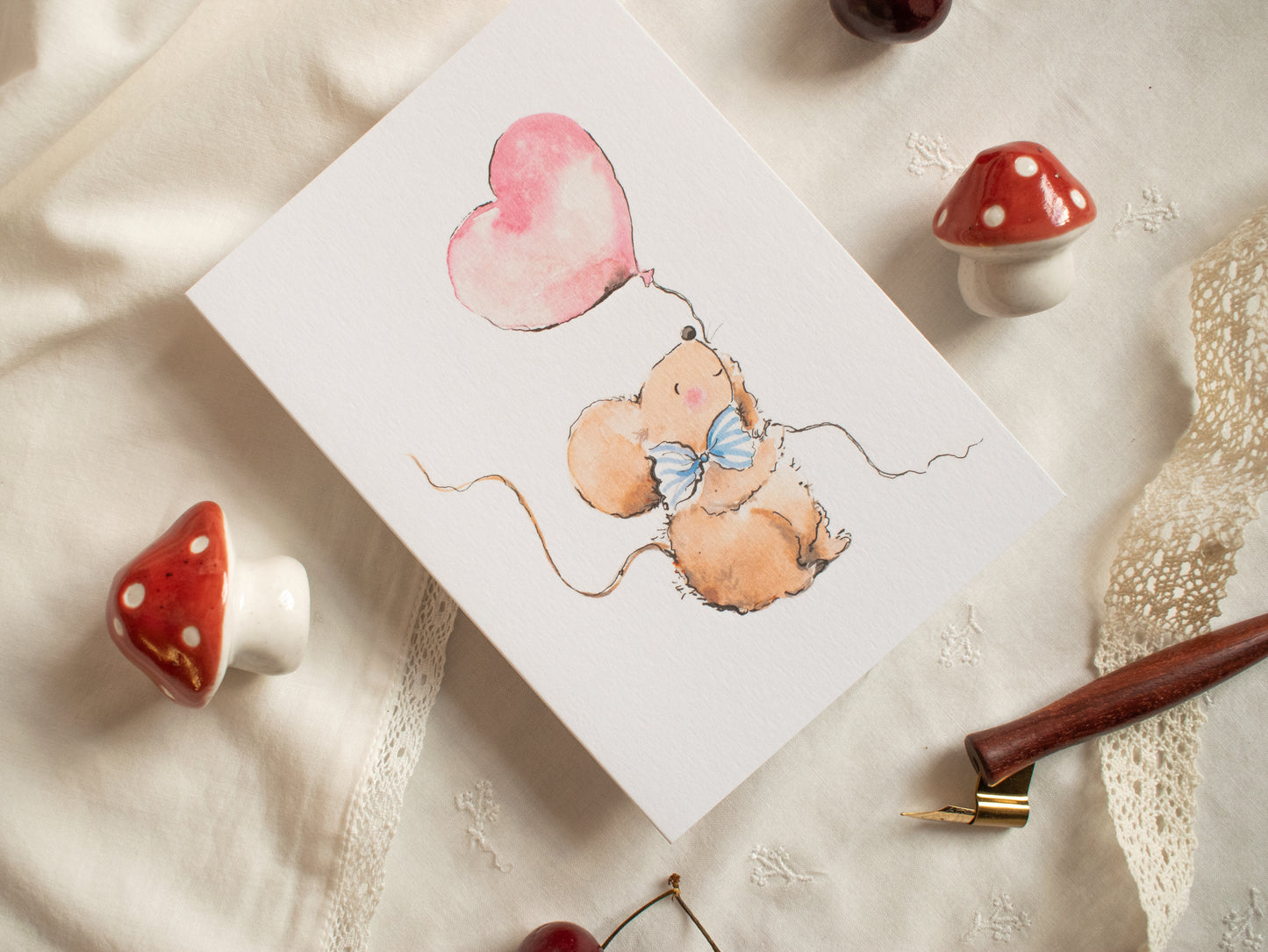 Card with a mouse holding a heart-shaped balloon on a textured surface with cherries and a pen.