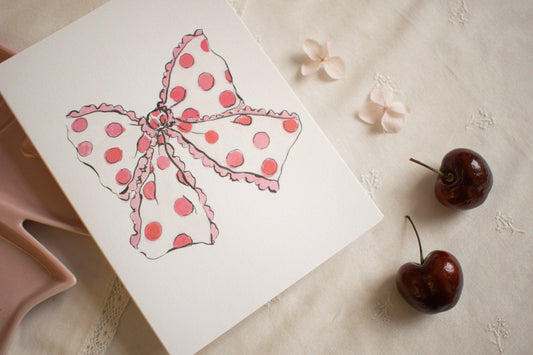 Card with a pink polka dot bow design, cherries, and flowers on a textured surface.