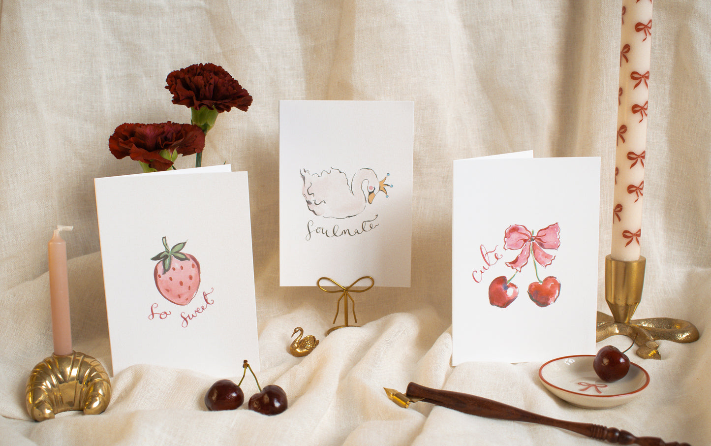 Three greeting cards with illustrations on a decorative table setting with candles and cherries.