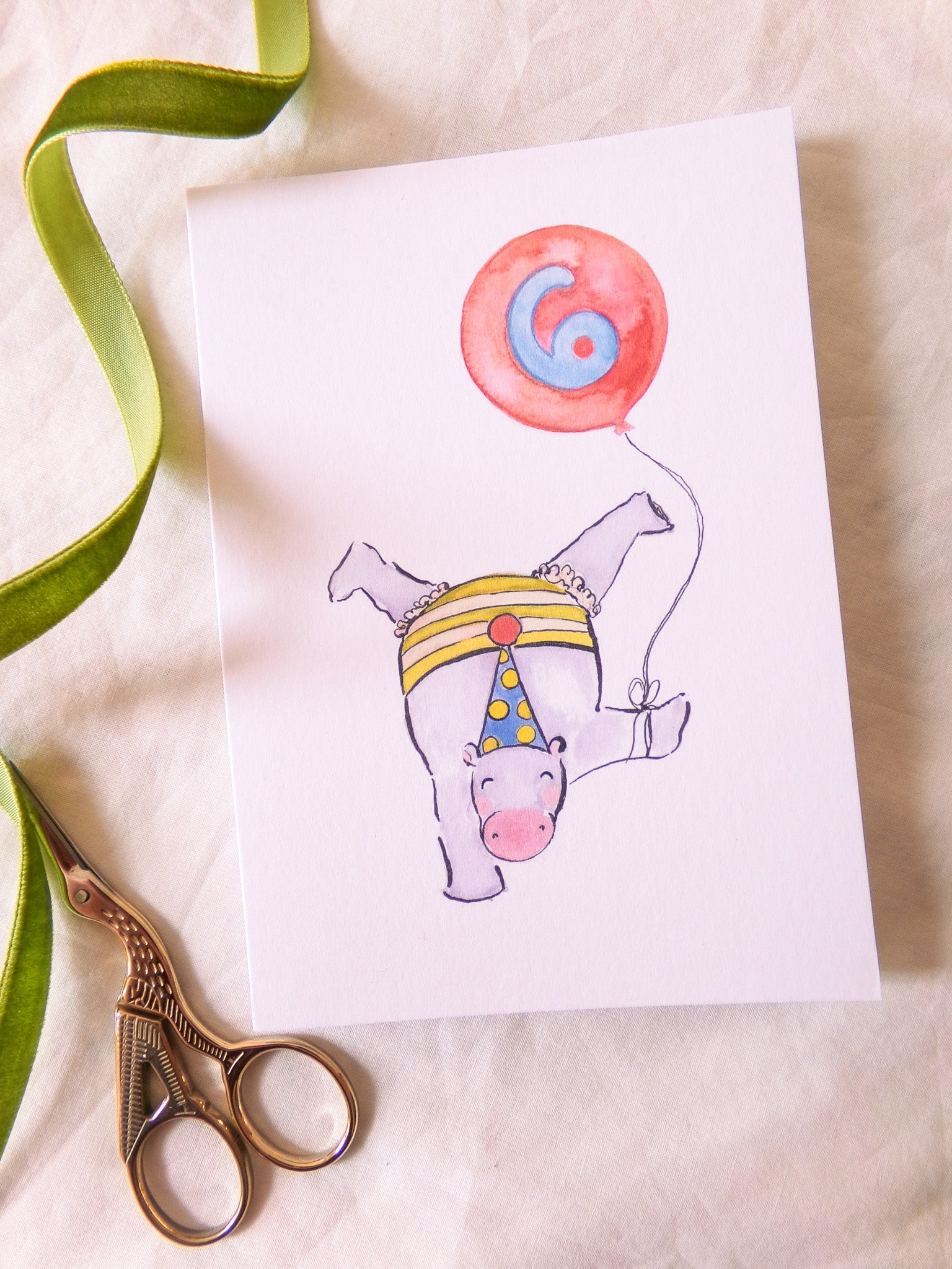 Illustration of a hippo with a party hat and balloon on a card, with scissors and ribbon on a beige background.