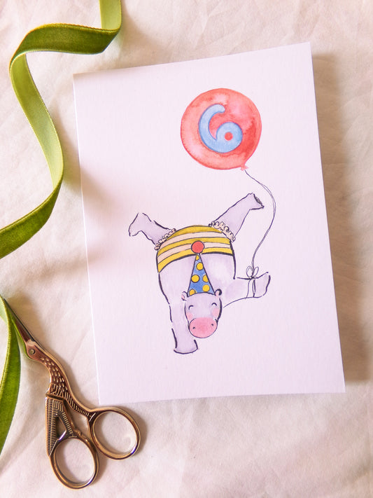 Illustration of a hippo with a party hat and balloon on a card, with scissors and ribbon on a beige background.