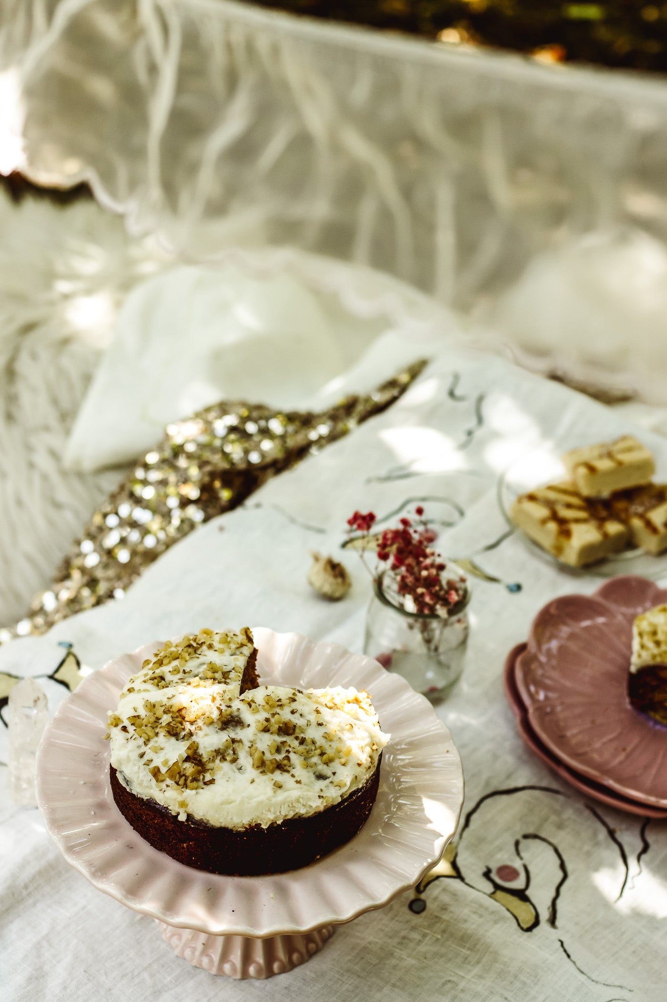 A heart-shaped chocolate cake with white frosting and chopped pistachio topping, served on a pink scalloped plate on a picnic blanket outdoors.