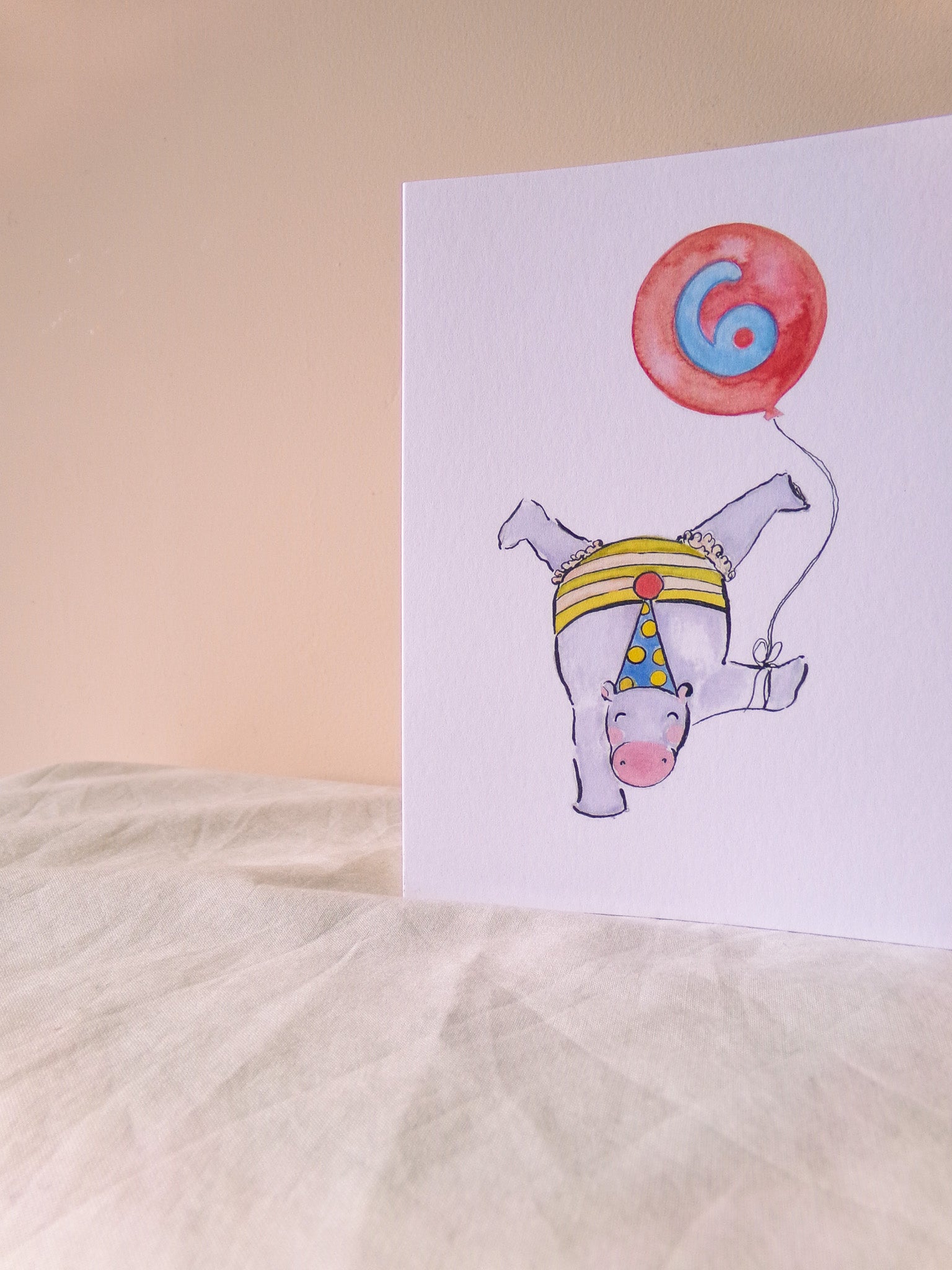 Card with a hippo wearing a party hat and holding a red balloon with the number 6 on a beige background.