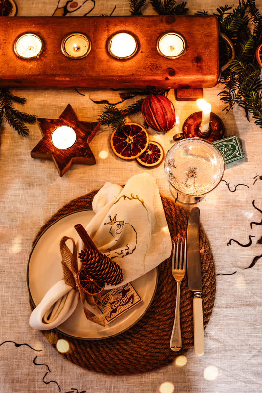 A styled holiday table setting with a place setting on a woven placemat, a glass of water, candles, dried orange slices, pinecones, and evergreen branches on a beige tablecloth.