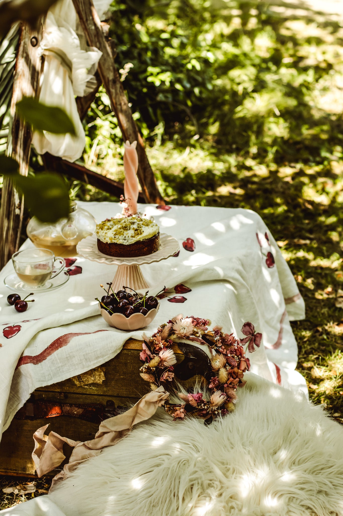 A styled outdoor picnic scene on a wooden crate with a white tablecloth, a small cake on a pedestal, a teacup, dried flowers, and a white faux-fur rug, set in a sun-dappled forest environment.
