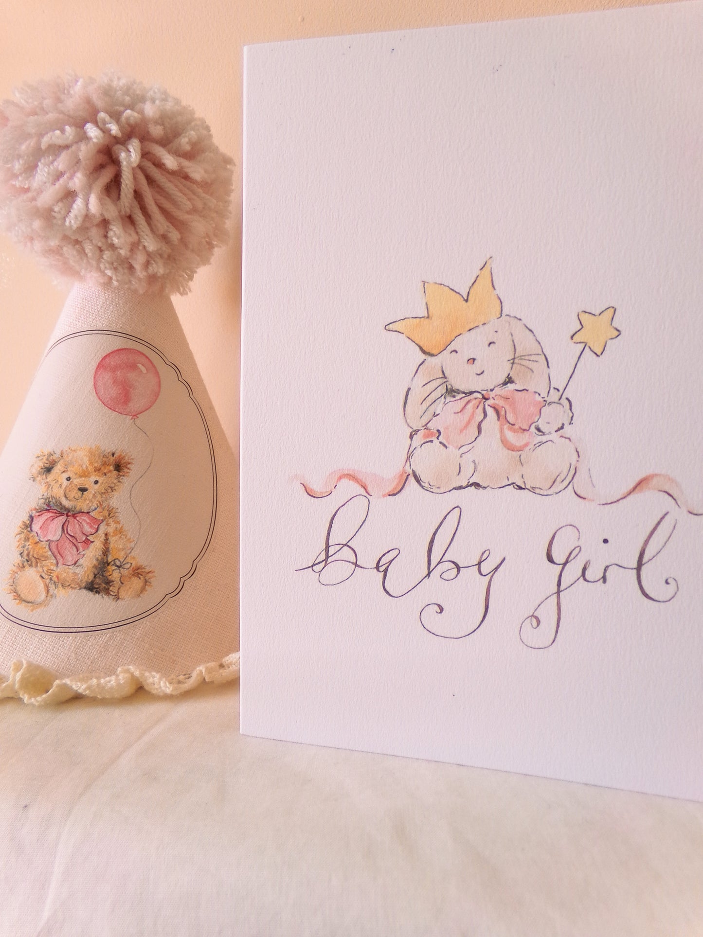 Baby girl-themed card with a teddy bear and crown design, next to a decorative hat with a pom-pom.