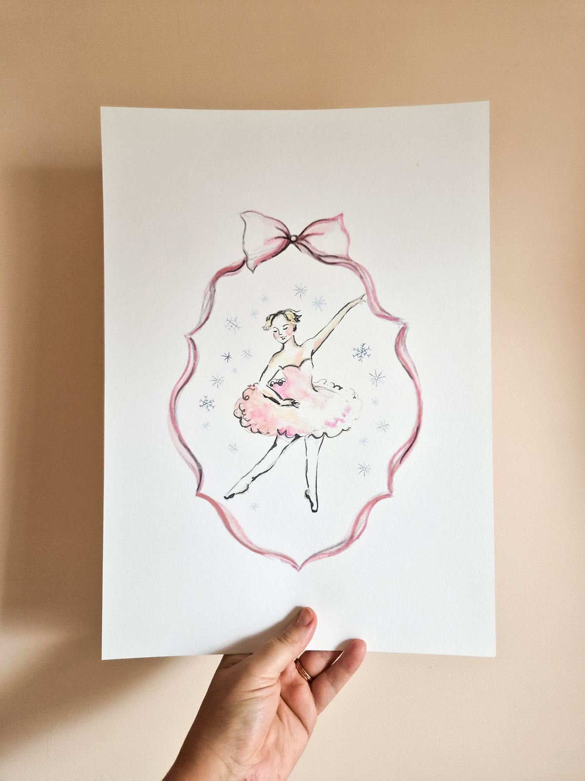 A hand holds a white art print featuring a watercolor illustration of a ballerina in a pink tutu and bow, set against a beige background.