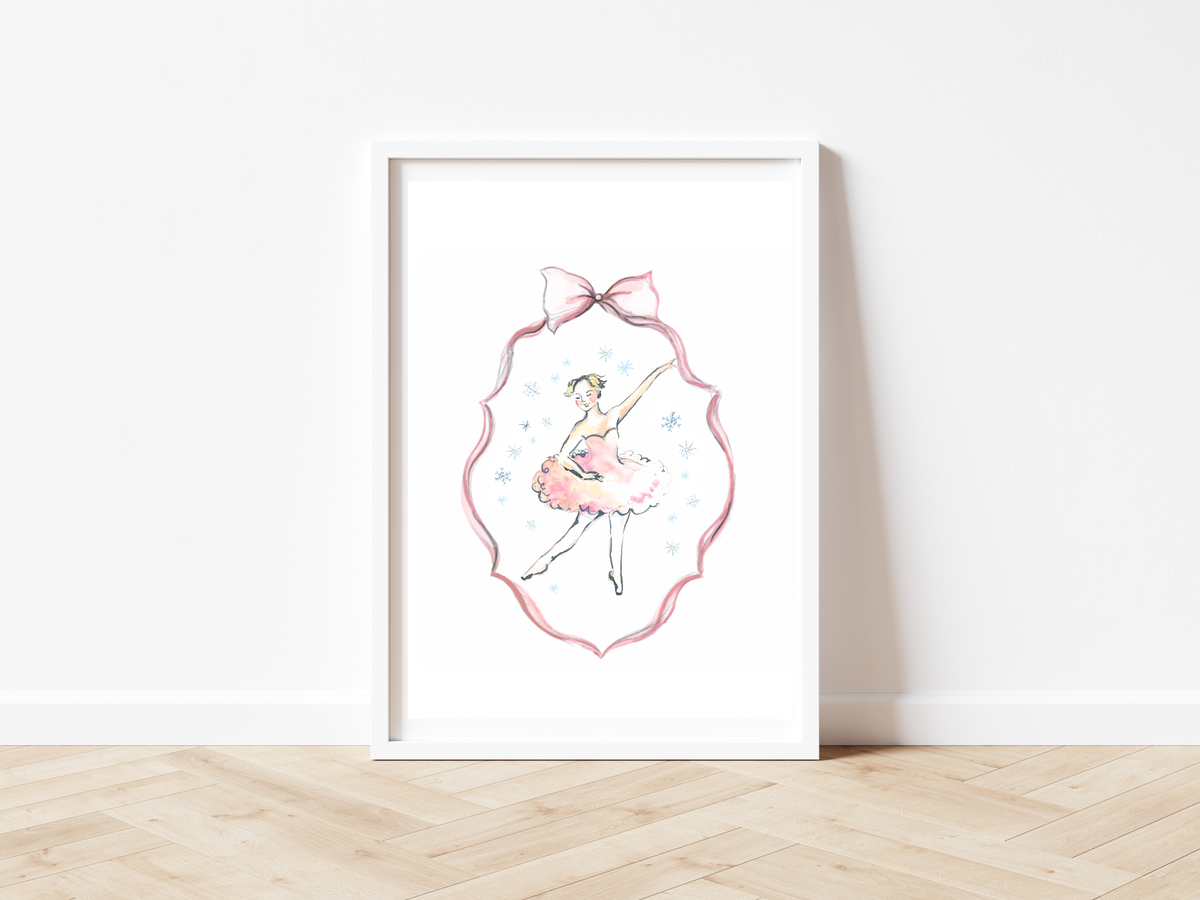 A framed art print featuring a watercolor ballerina in a pink tutu and bow, displayed in a white frame leaning against a white wall on a light wood floor.