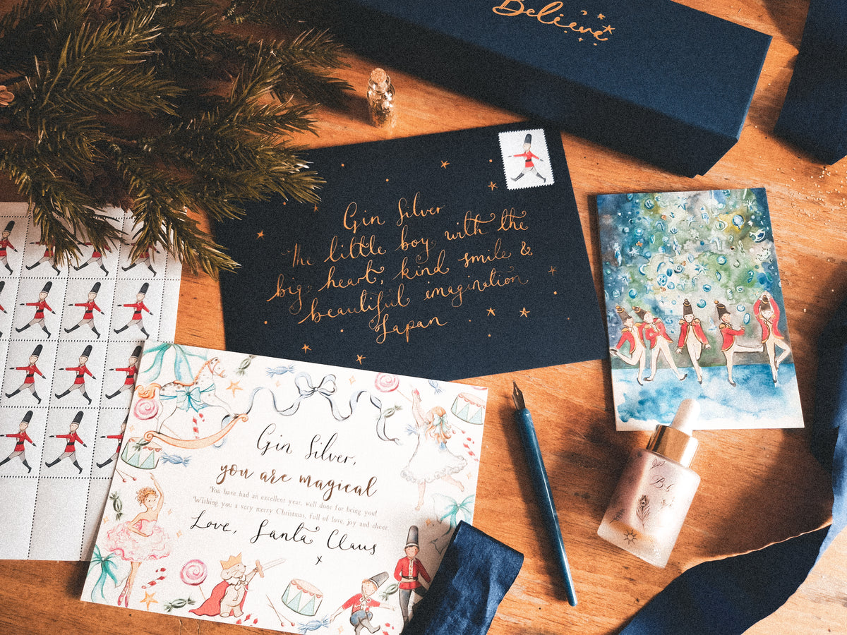 A styled flat-lay of a Gin Filler stationery set on a wooden table, featuring a navy blue box, a navy blue card with gold foil writing, a white card with colorful illustrations, a sheet of red-and-black toy soldier stickers, a watercolor card, and a small bottle.