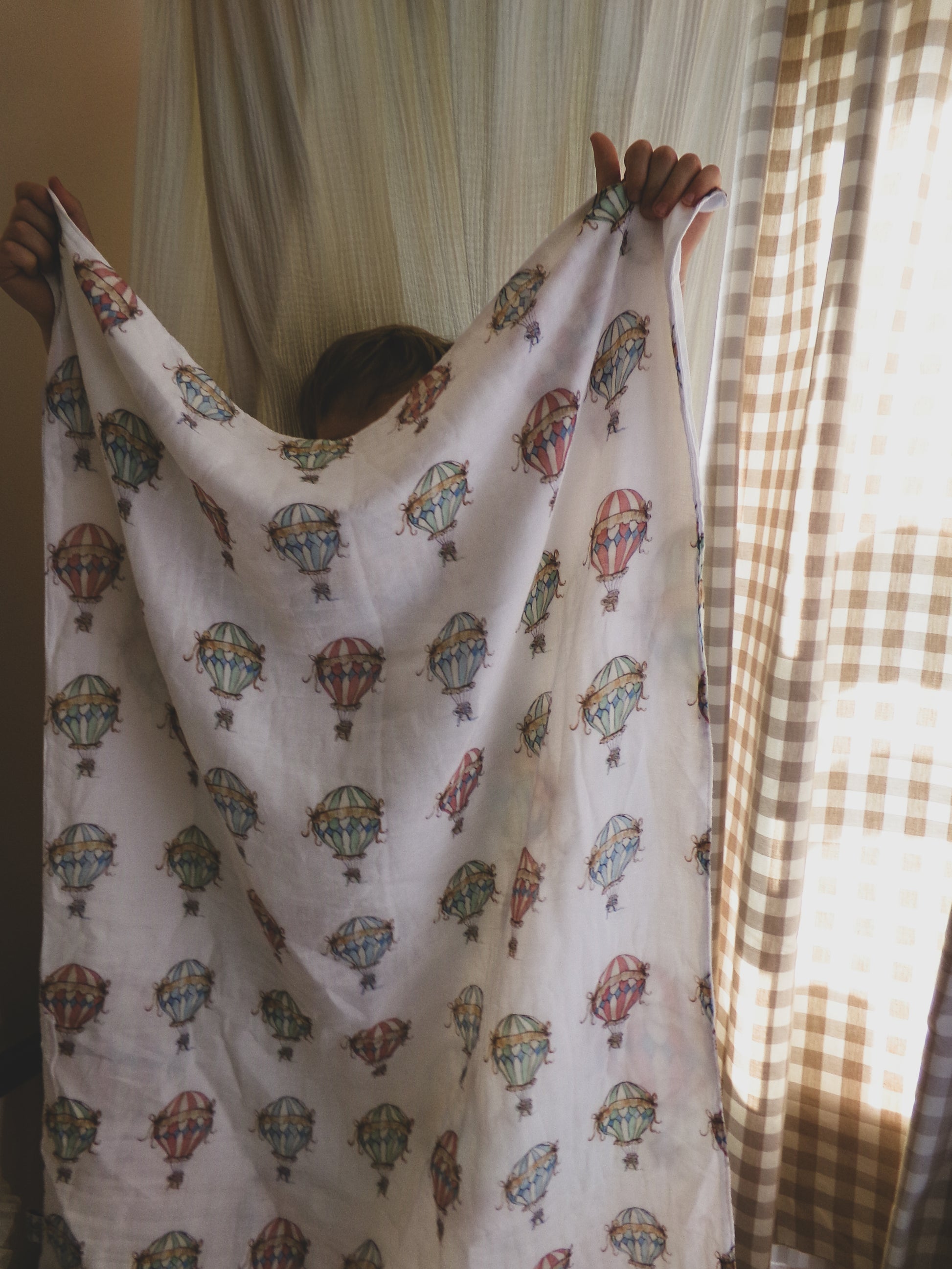 Person holding a white fabric with colorful hot air balloon patterns in front of a window with curtains.