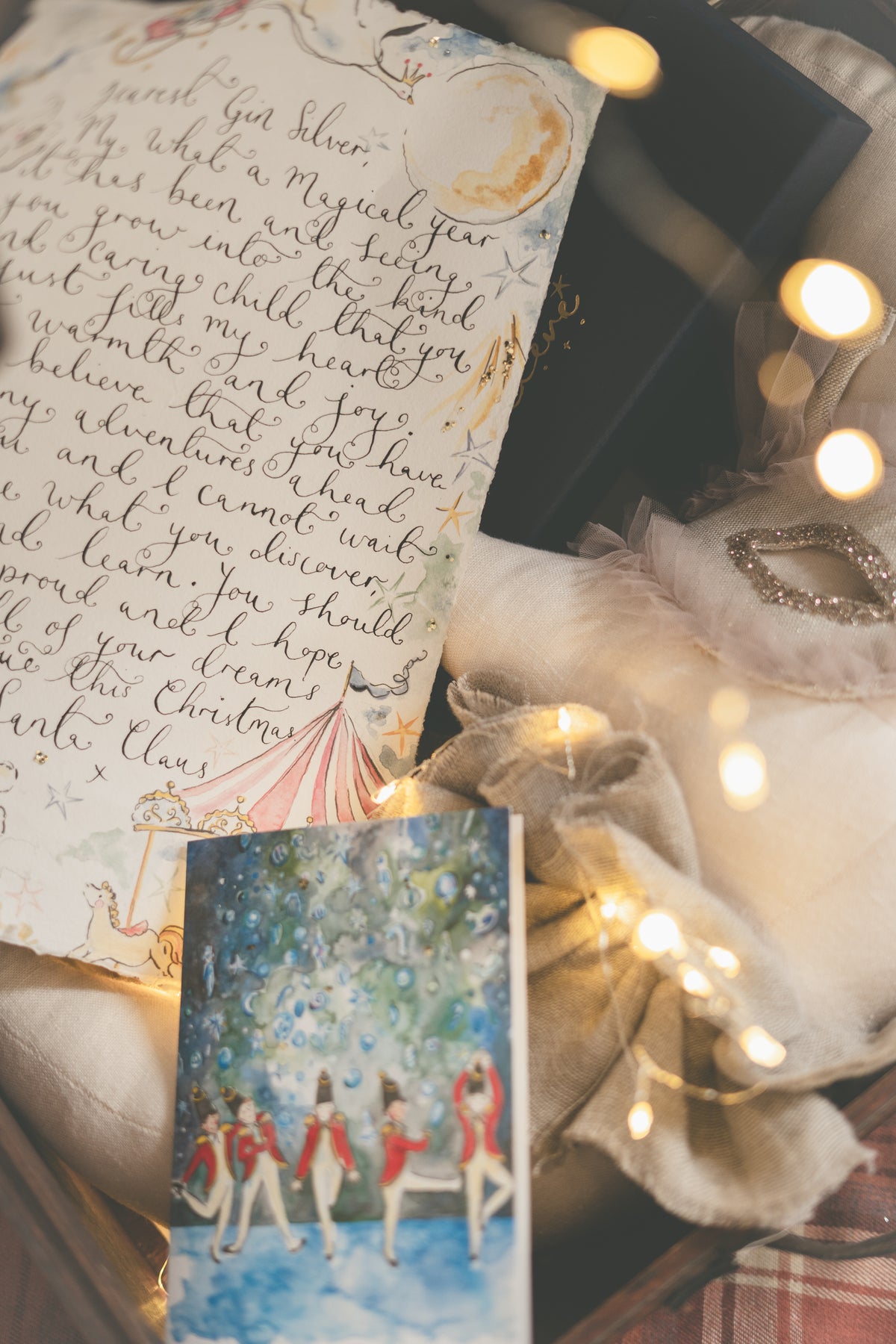 A styled flat-lay of a Christmas-themed card featuring an illustration of children in red and white outfits under a starry night sky, surrounded by warm fairy lights, a beige scarf, a black gift box, and a piece of handwritten paper.