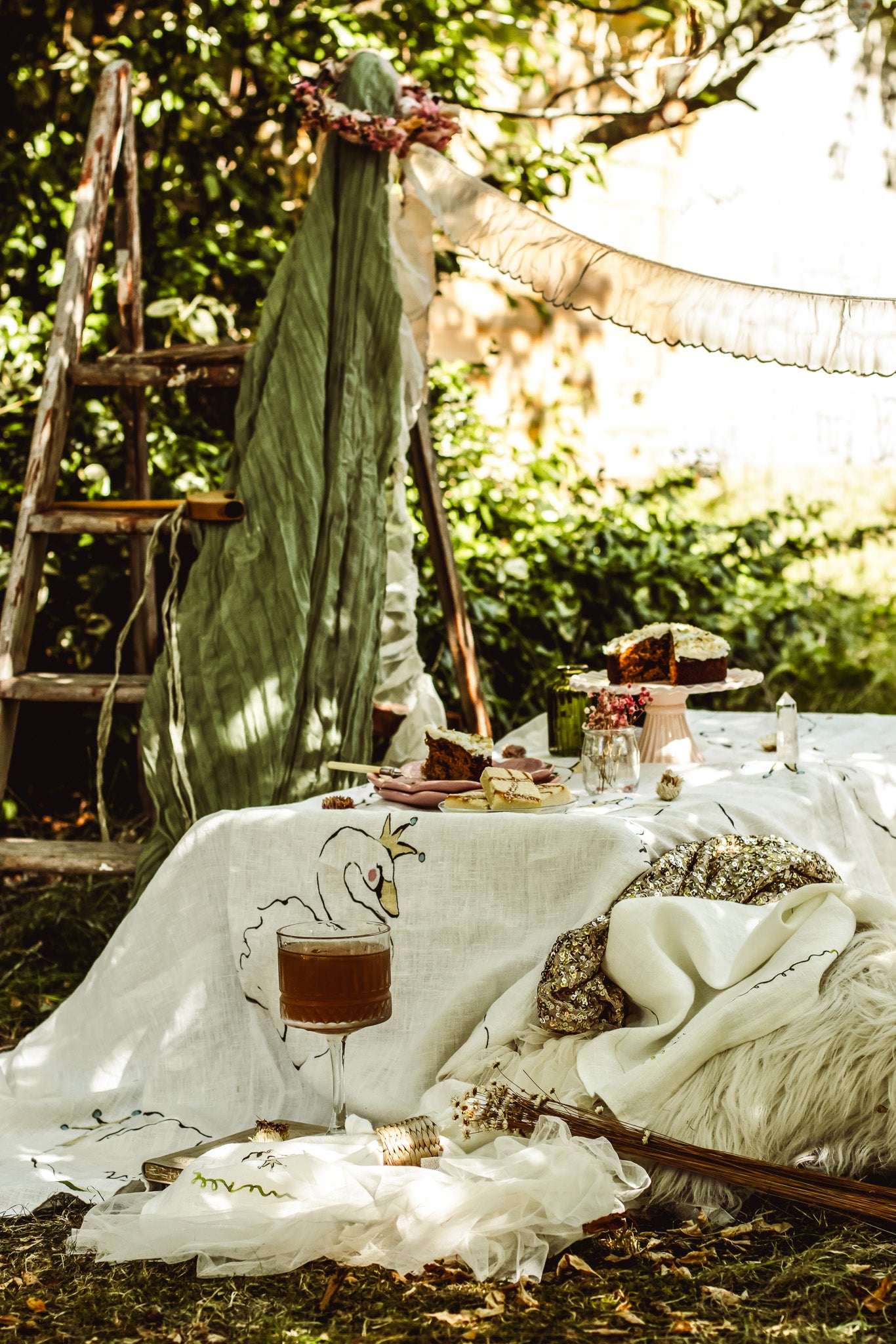 A rustic outdoor picnic setup under a tree canopy, featuring a white embroidered tablecloth, a green draped fabric backdrop, a glass of iced coffee, and a blanket with embroidered details.