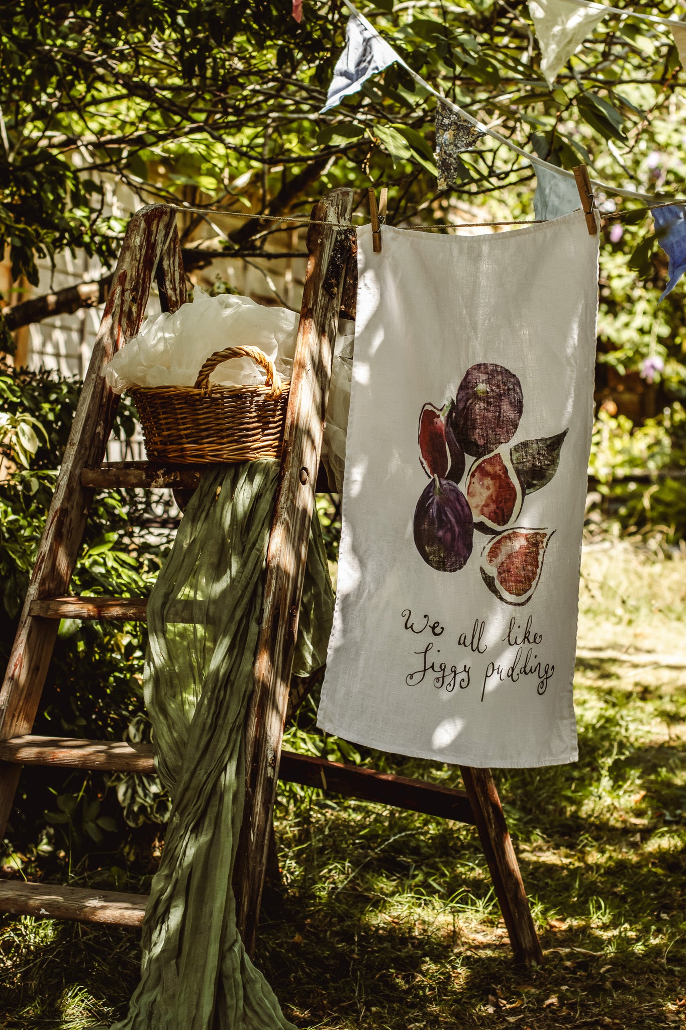 A white tea towel with a watercolor print of figs and the text 'we all like figs pretty' hangs on a rustic wooden ladder outdoors in a garden setting.