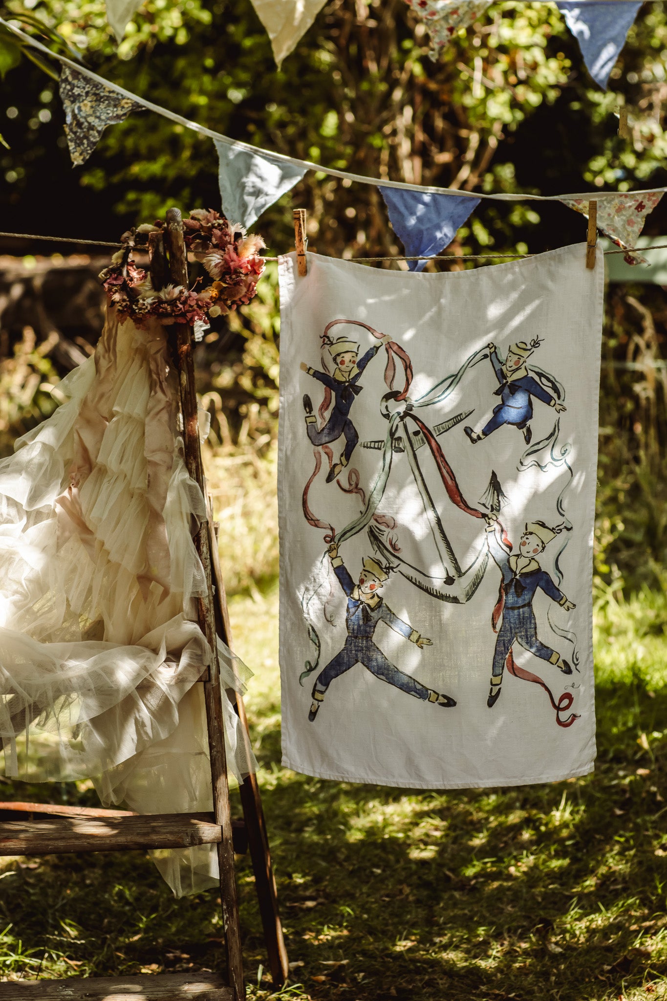 A white fabric banner or tapestry featuring vintage illustrations of children in blue and red outfits, hanging outdoors on a clothesline next to a rustic wooden chair with a cream dress.