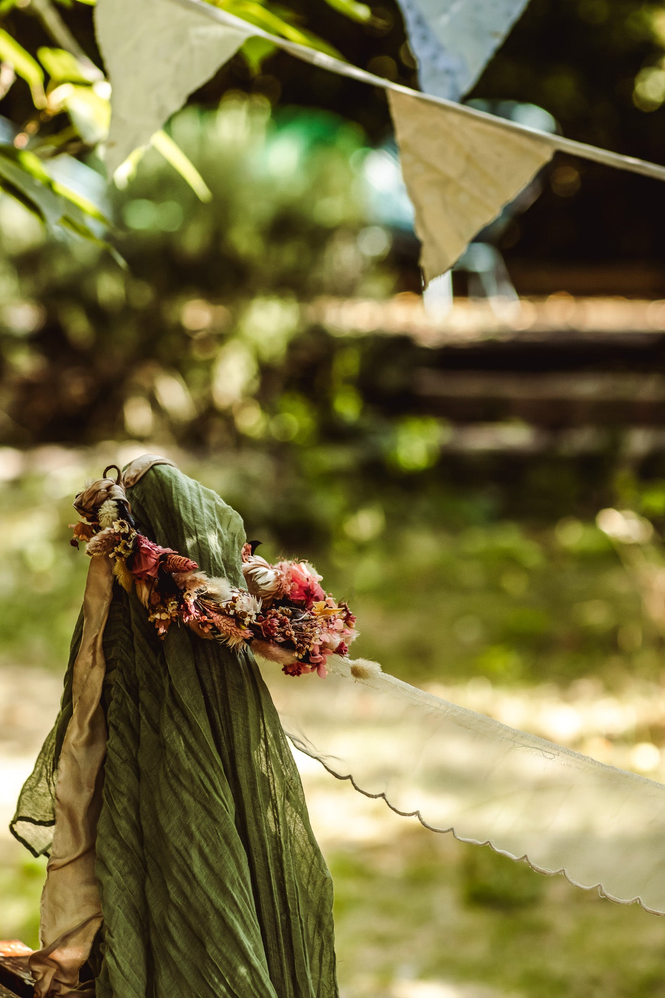 A close-up of a green fabric item, possibly a scarf or shawl, adorned with a floral crown and hanging from a string in an outdoor garden setting.