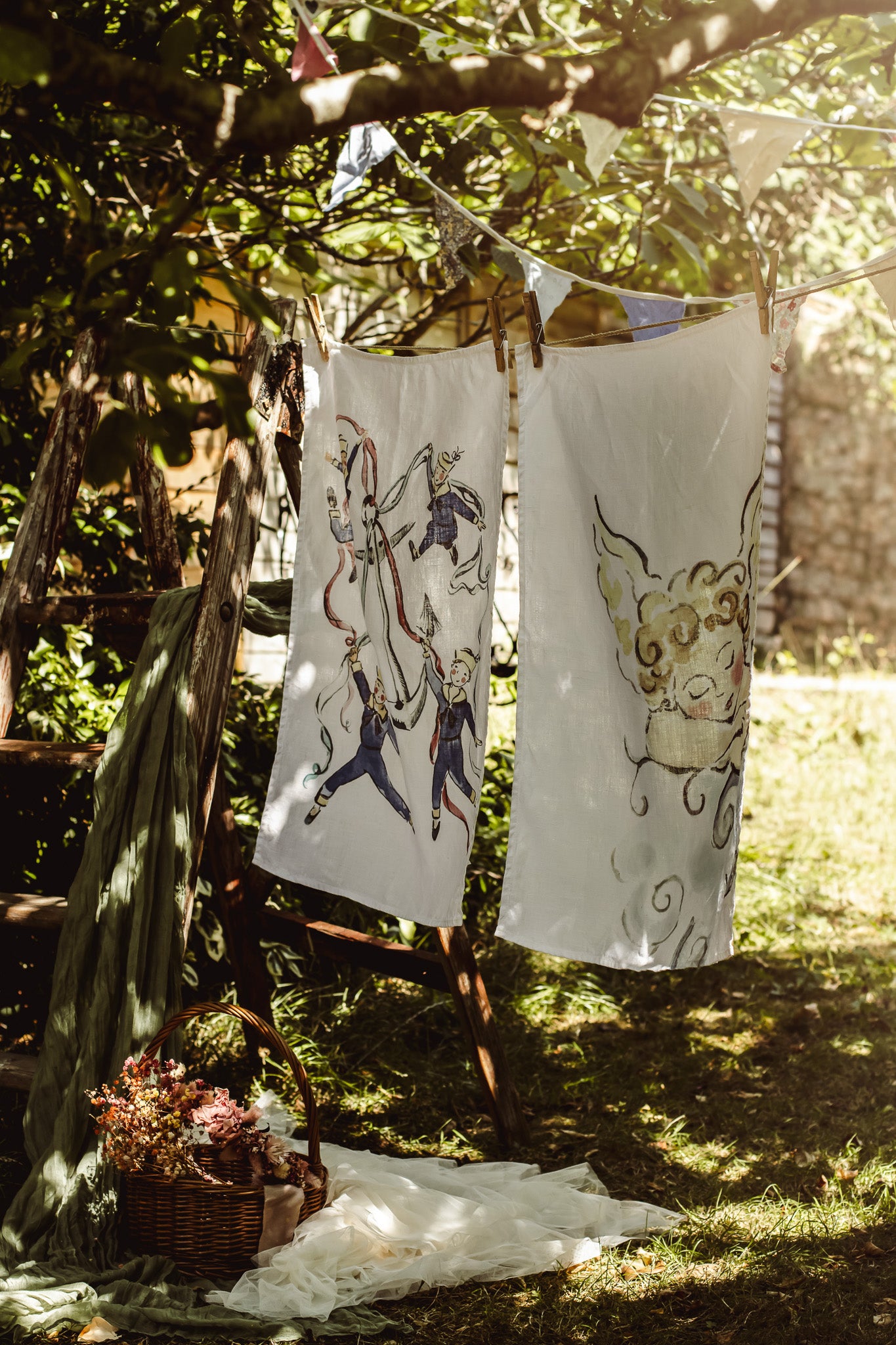 Two white fabric cloths or napkins with hand-drawn whimsical illustrations hang on a string outdoors under a tree, with sunlight filtering through the leaves and a basket of flowers on the ground below.