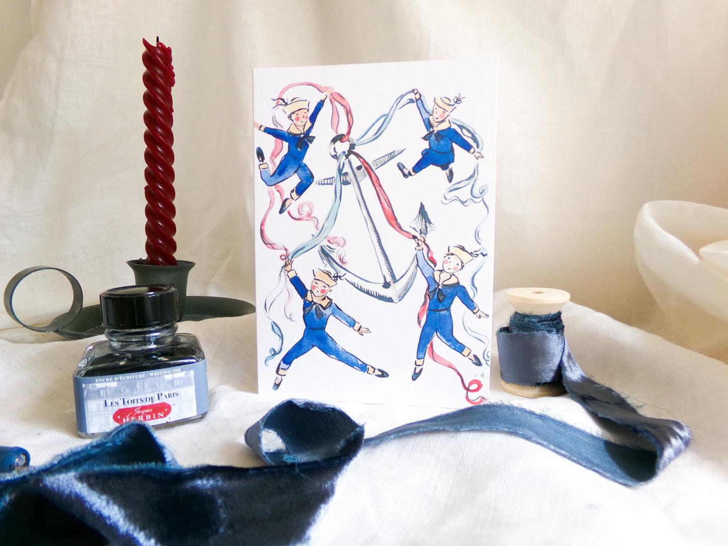 A white greeting card featuring a whimsical illustration of four children in blue outfits playing with long red and blue ribbons, styled on a draped cream fabric background with sewing and craft props including a red spiral candle, ink bottle, ribbon, and thread spool.