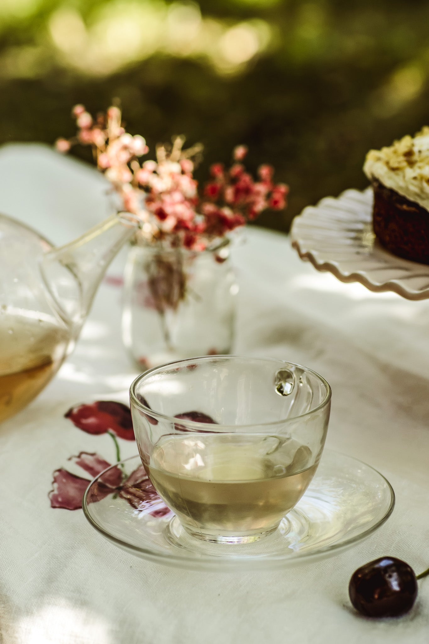 A clear glass teacup and saucer filled with tea, set on a white tablecloth outdoors with a blurred green garden background. A glass teapot, dried pink flowers in a vase, and part of a cupcake are also visible.