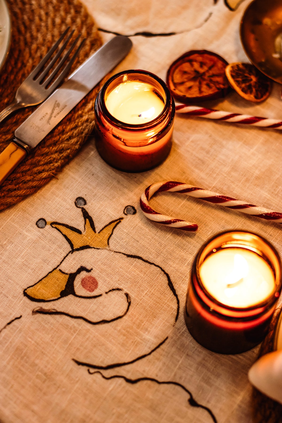 A cozy tabletop scene with two lit amber glass candles on a beige fabric runner printed with a stylized bird illustration. Candy canes, dried orange slices, a fork, knife, and a woven placemat are arranged around the candles.