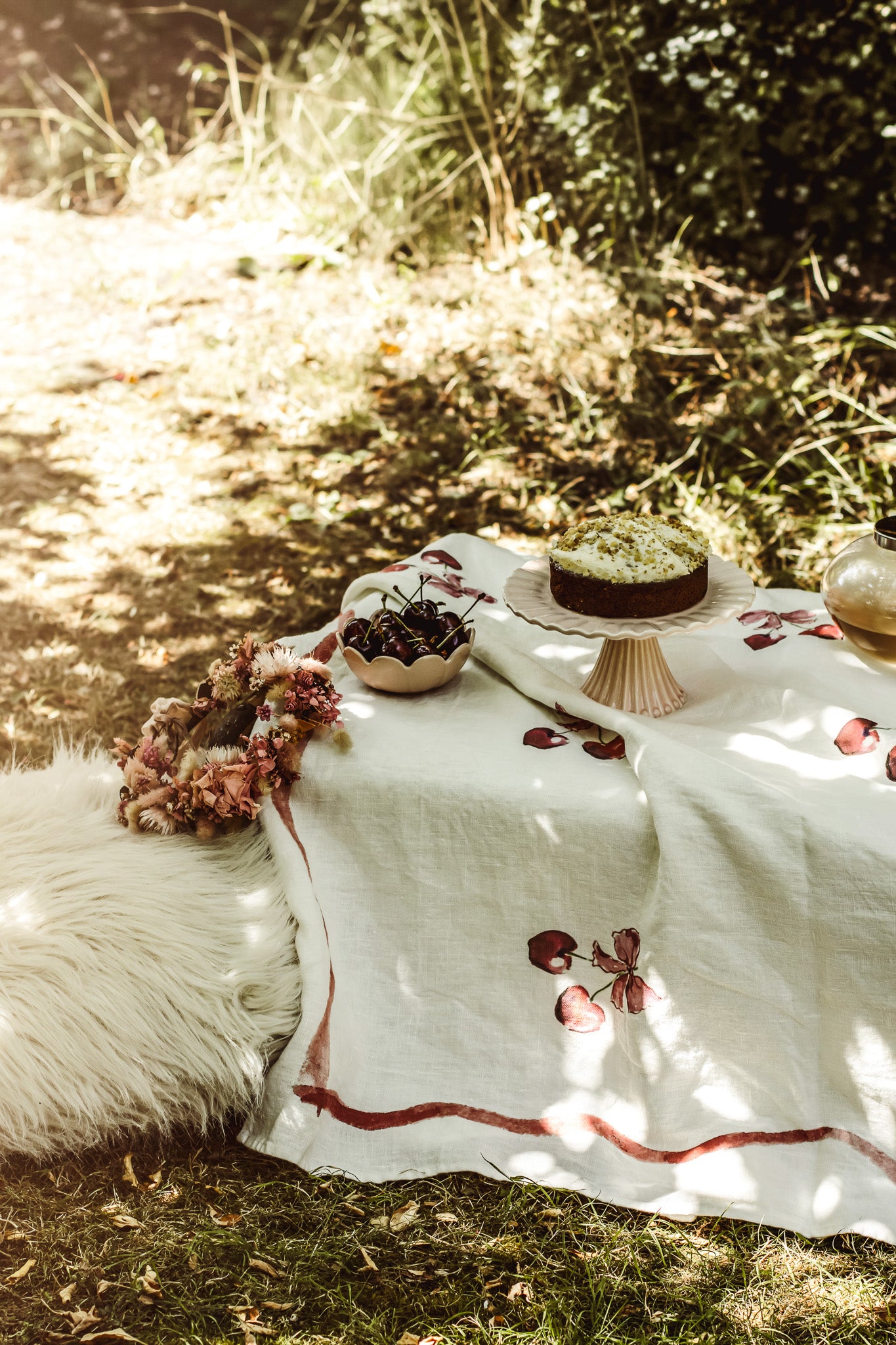 An off-white linen picnic blanket with red embroidered floral details is spread on grass in a sunlit outdoor setting. A small cake on a stand, a bowl of dark berries, and dried flowers are arranged on the blanket.