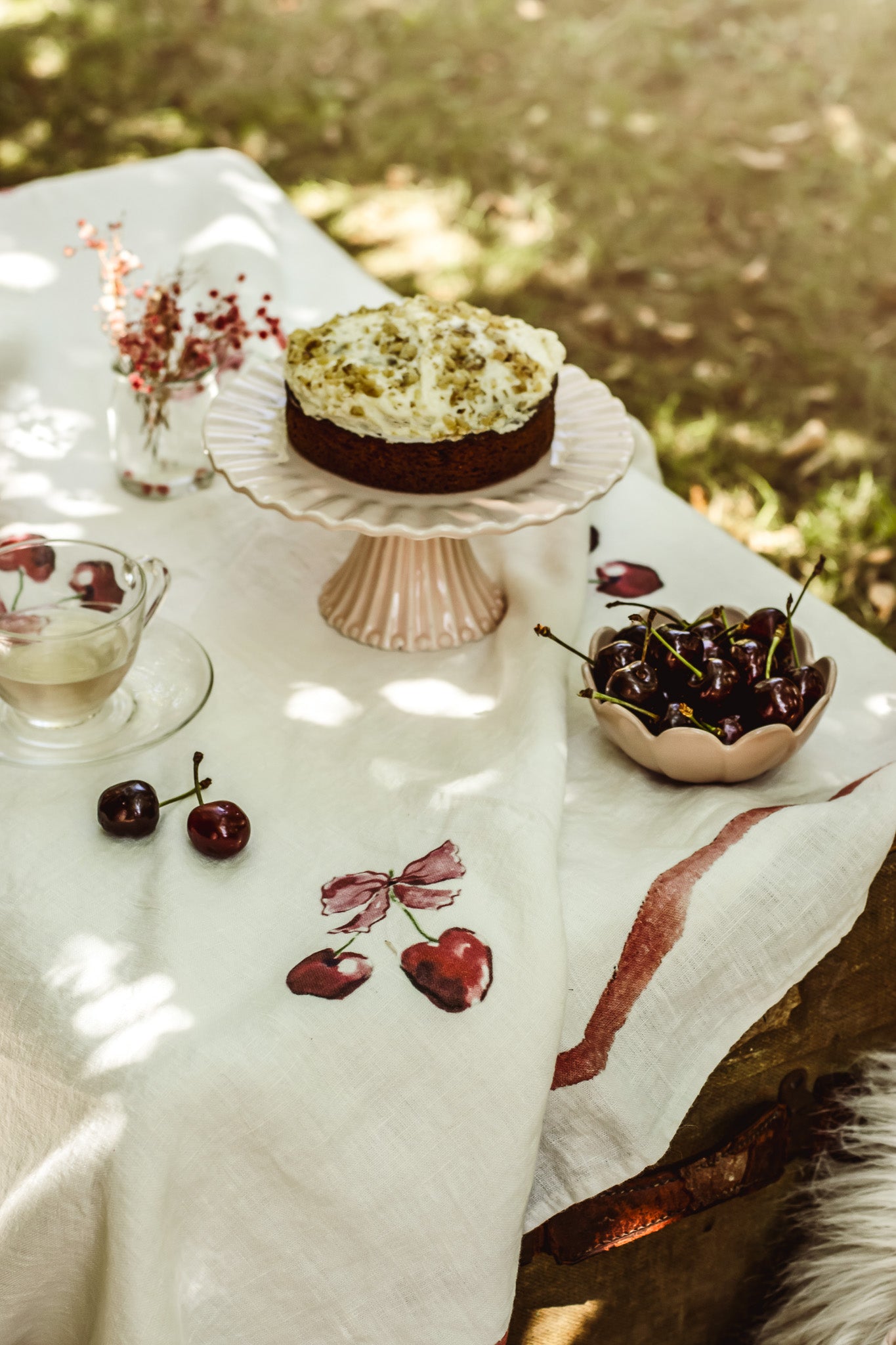 A styled outdoor table scene with a cherry-themed dessert on a pedestal stand, a bowl of fresh cherries, scattered cherries on a white embroidered cloth, and a glass cup in a sunlit garden setting.