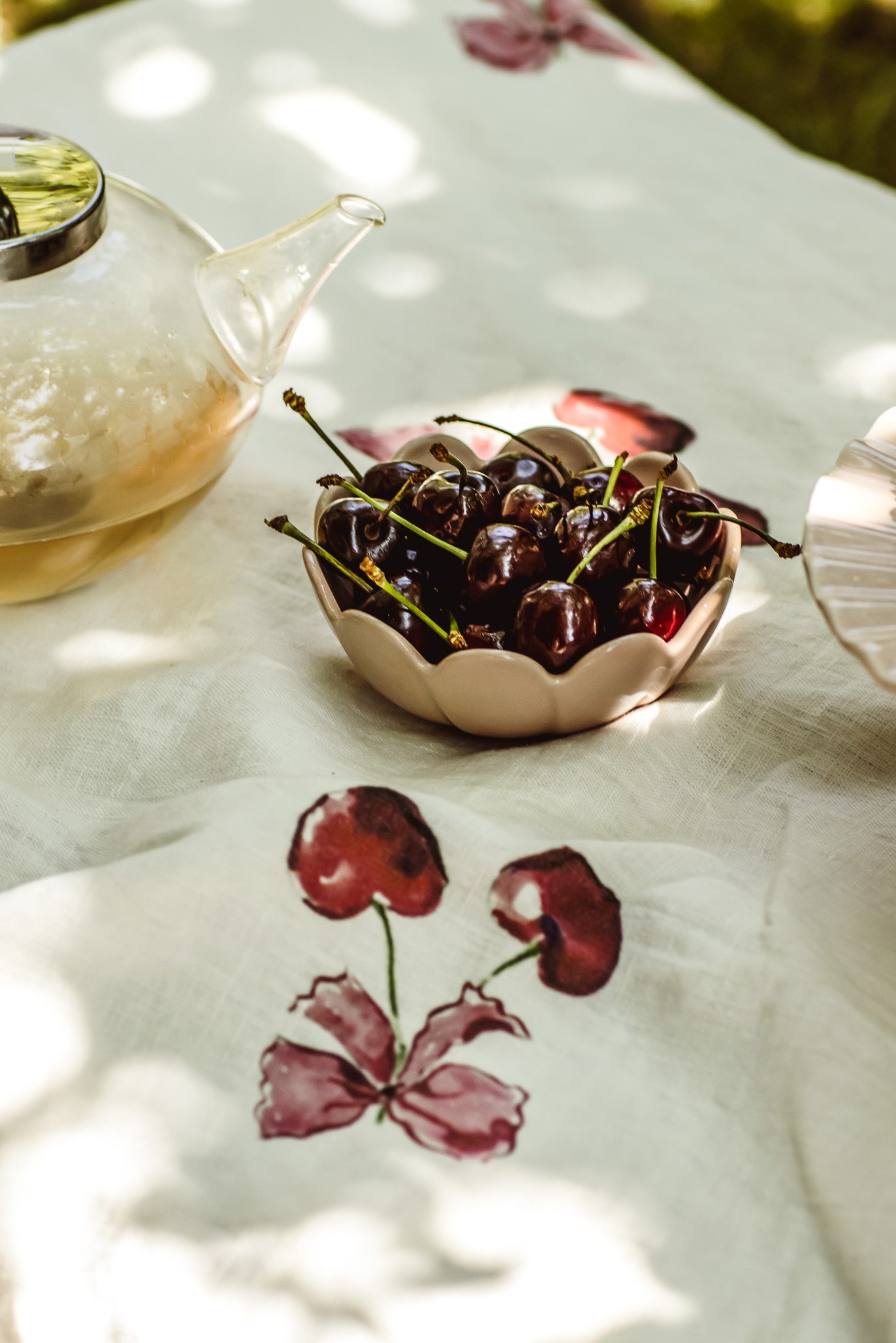 A ceramic bowl filled with dark cherries sits on a white tablecloth printed with red cherries, next to a glass teapot in dappled sunlight.