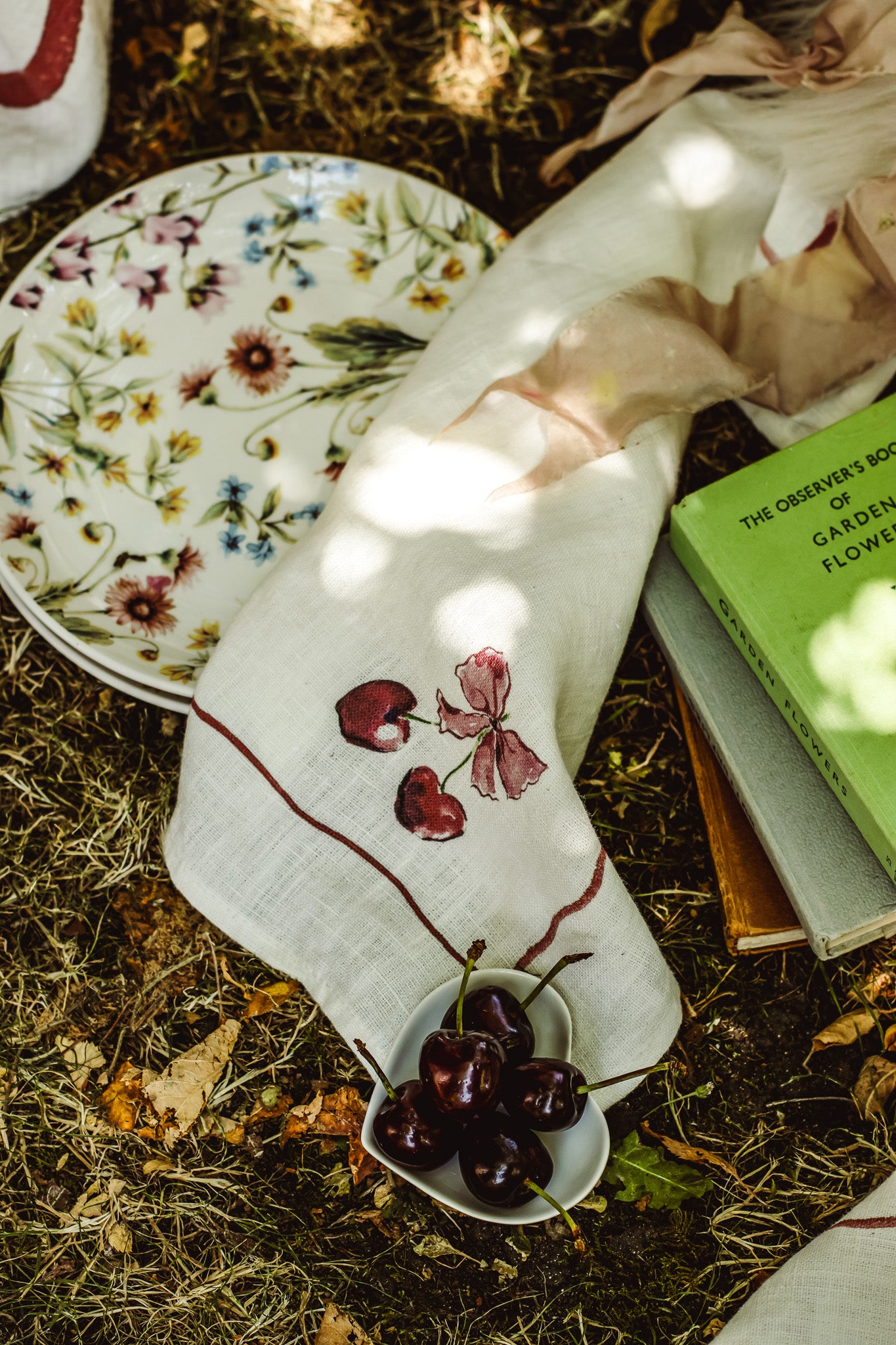 A styled flat-lay of a white linen napkin with embroidered cherries and a floral plate on a forest floor background.