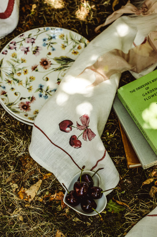 A styled flat-lay of a white linen napkin with embroidered cherries and a floral plate on a forest floor background.