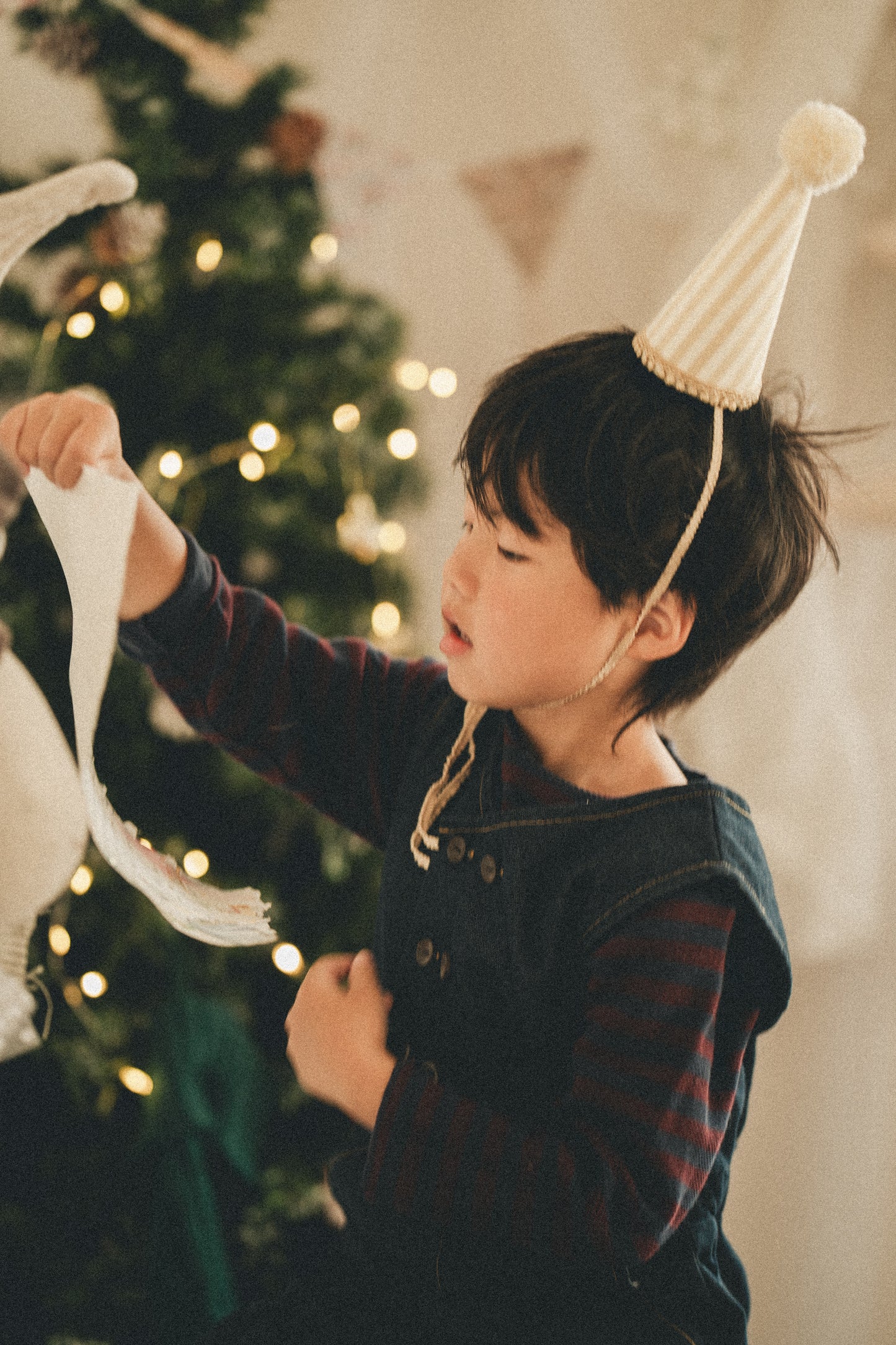 A young boy wearing a striped party hat and dark overalls with red striped sleeves, holding a white ornament in front of a softly lit Christmas tree.
