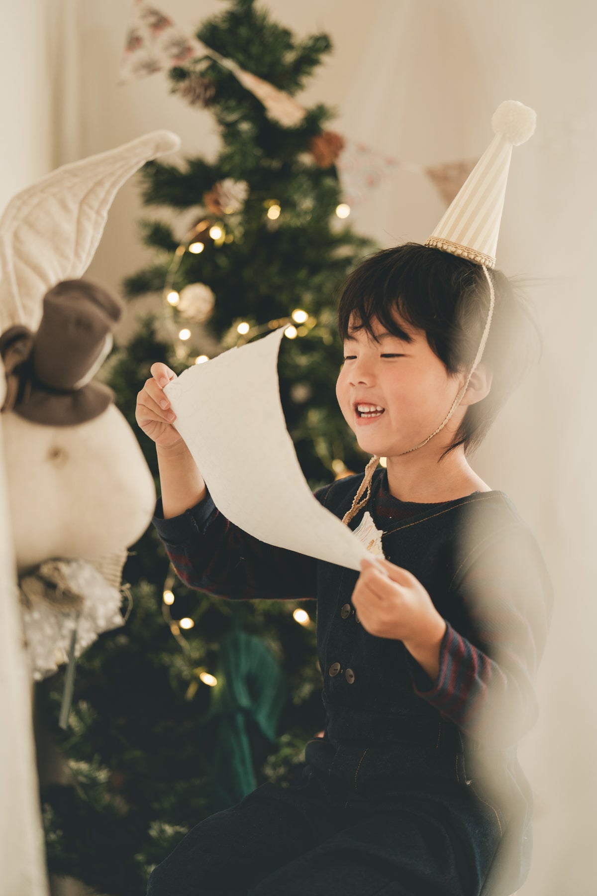 A young child wearing a white party hat and dark clothing holds a piece of paper in front of a softly blurred Christmas tree with warm lights.