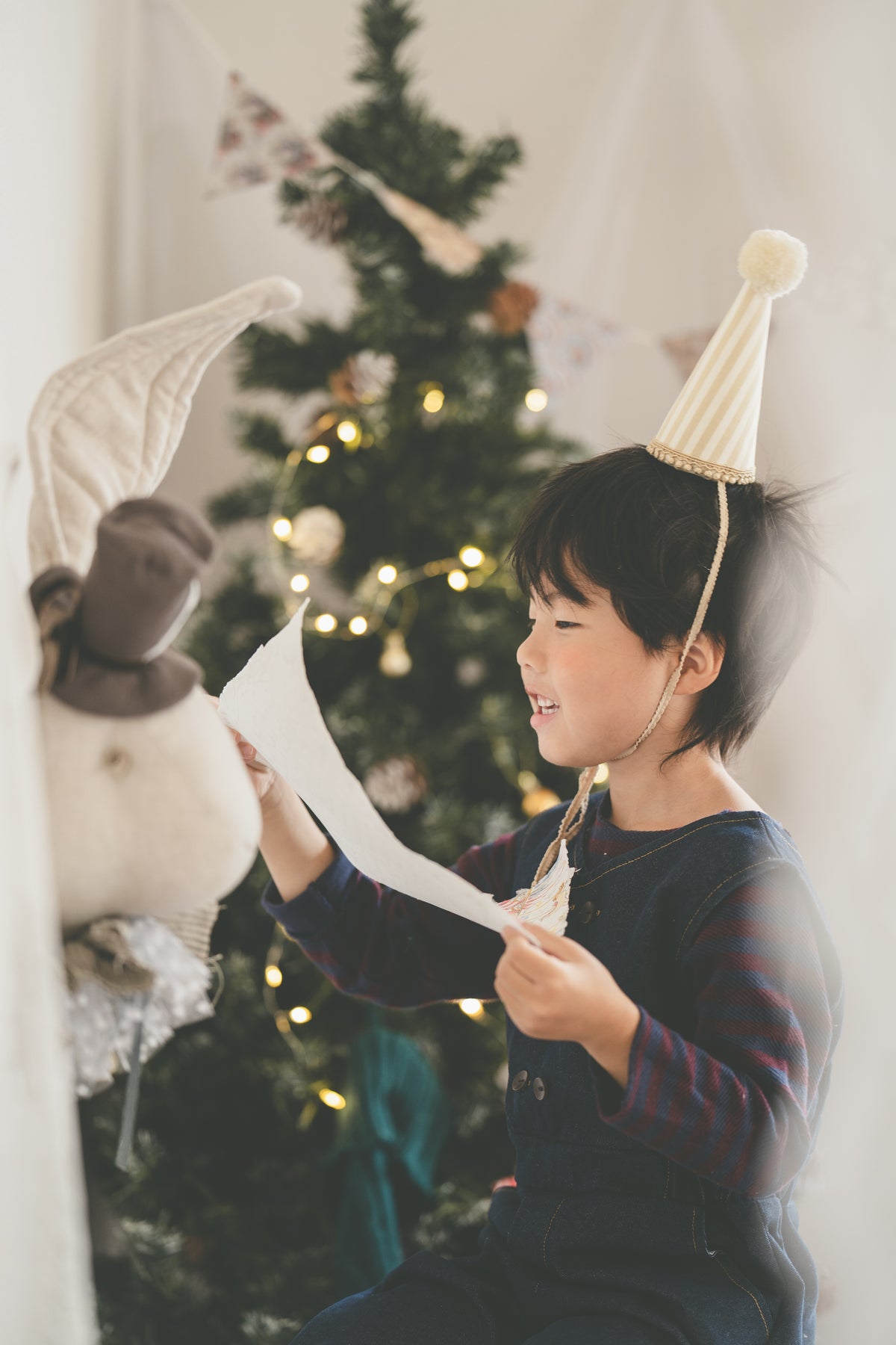 A young child wearing a striped party hat and dark overalls sits in front of a decorated Christmas tree, holding a piece of paper. A plush toy is visible on the left side of the frame.