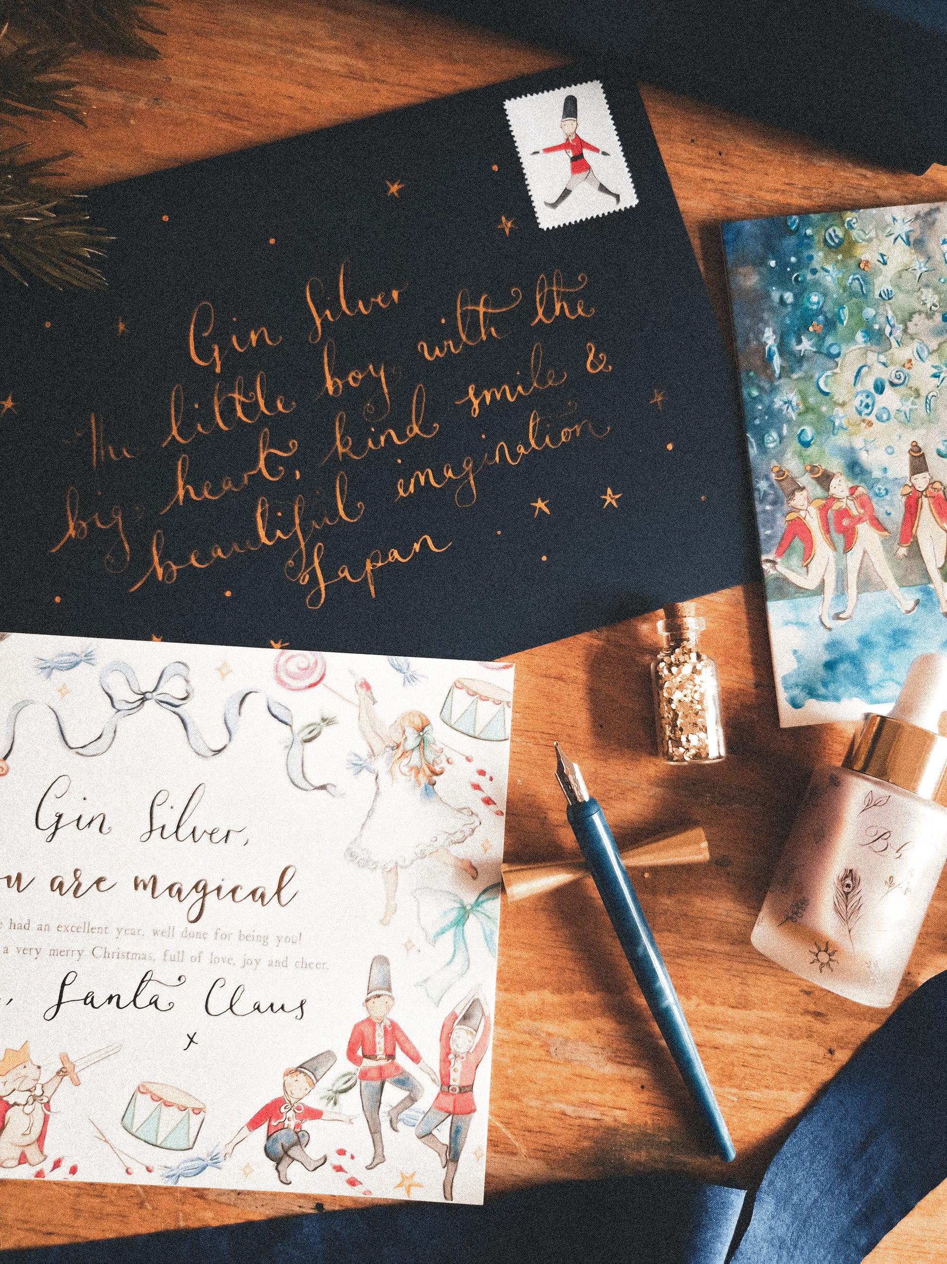 A styled flat-lay of printed cards and stationery on a wooden surface, including a dark card with gold cursive text and a white illustrated card with Santa and ballet dancers, alongside a blue pen, glitter bottle, and other decorative items.