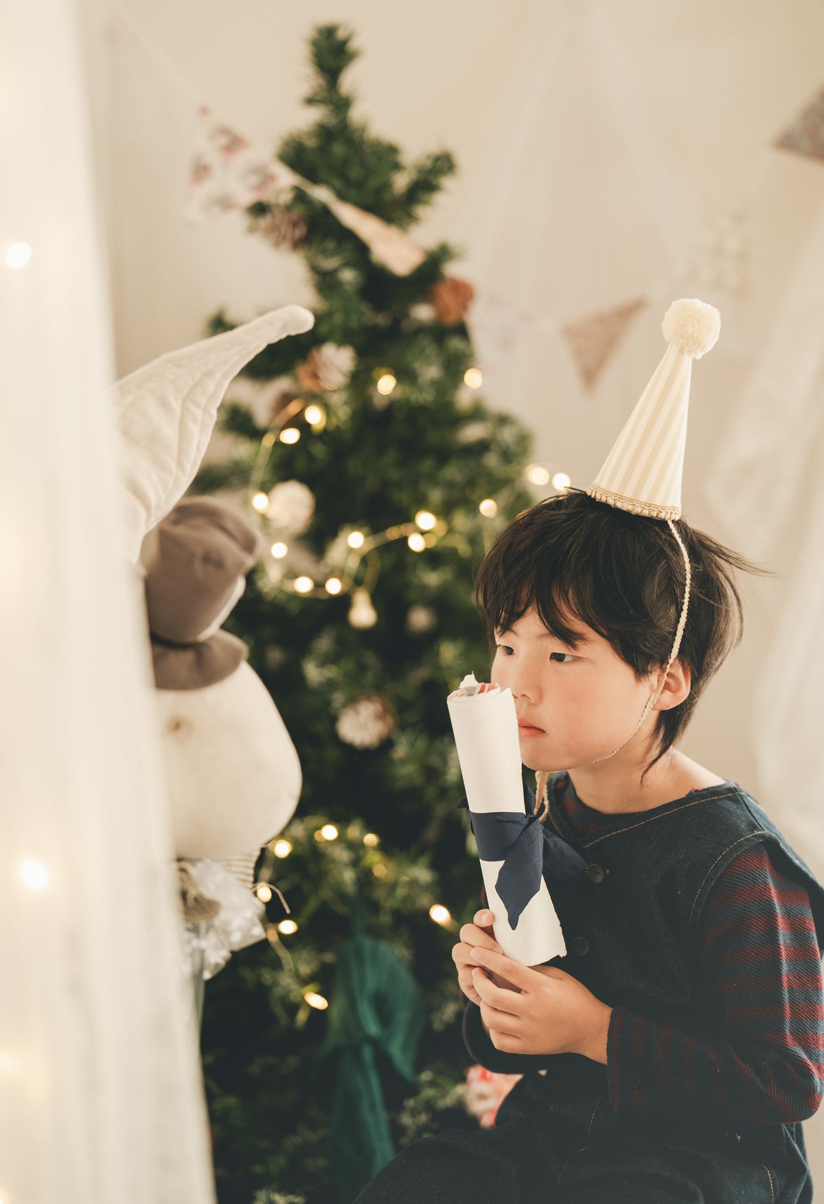A young boy wearing a white party hat and a dark striped shirt sits in front of a decorated Christmas tree, holding a rolled white paper scroll with a dark ribbon.