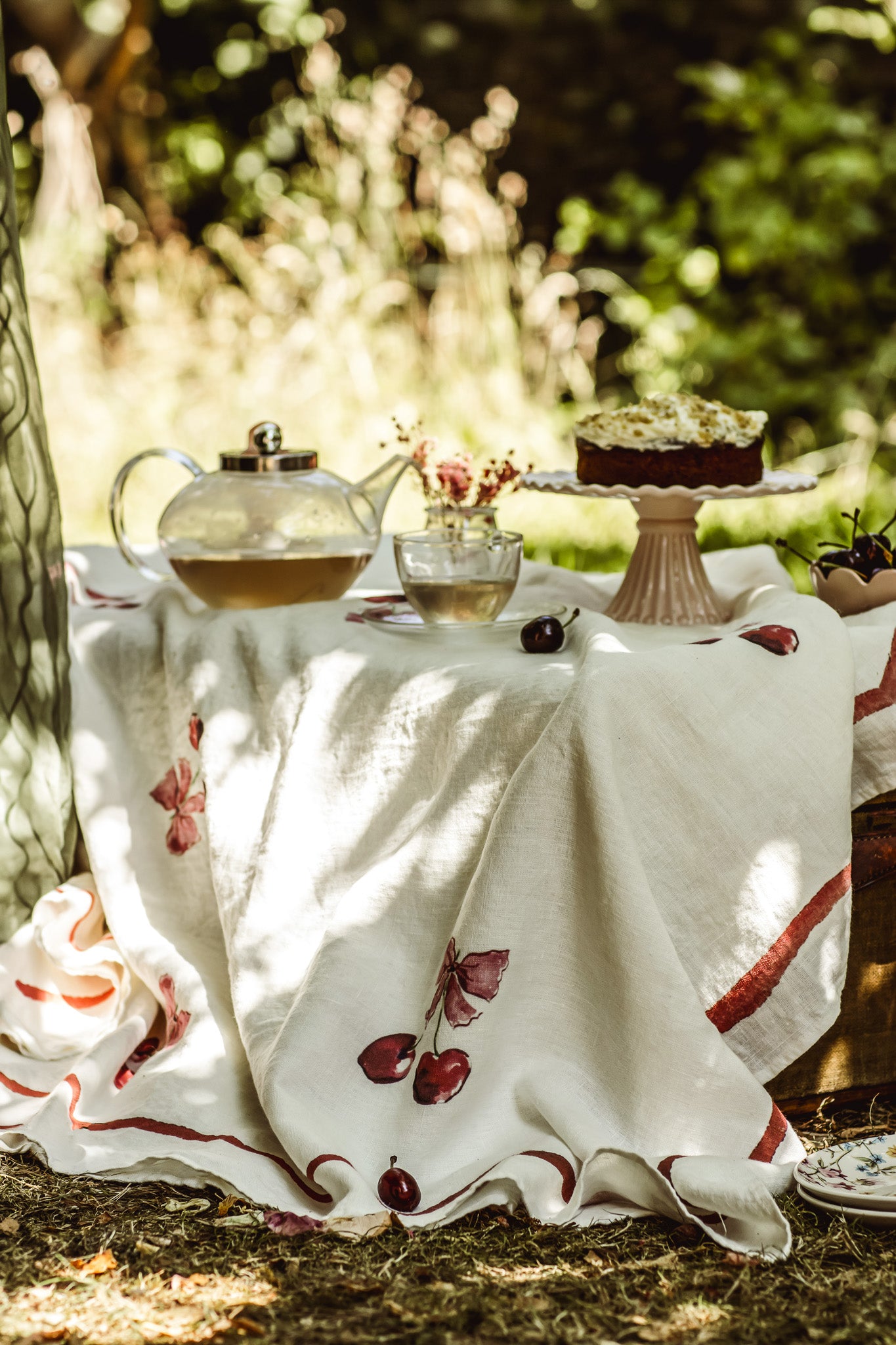 A white linen tablecloth with red cherry and stripe patterns draped over a picnic table outdoors, styled with a glass teapot, a small vase of flowers, and a cake on a pedestal stand.