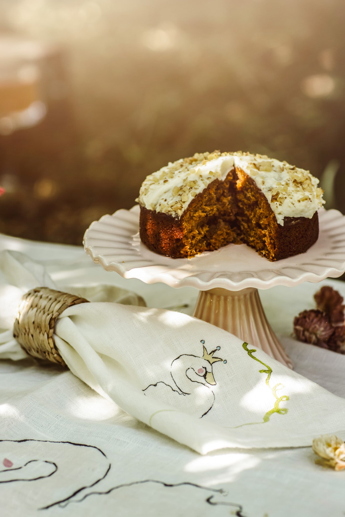A slice of carrot cake with white frosting sits on a white pedestal stand, set on a table with a white napkin, a woven napkin ring, and pinecones in a warm, sunlit outdoor setting.