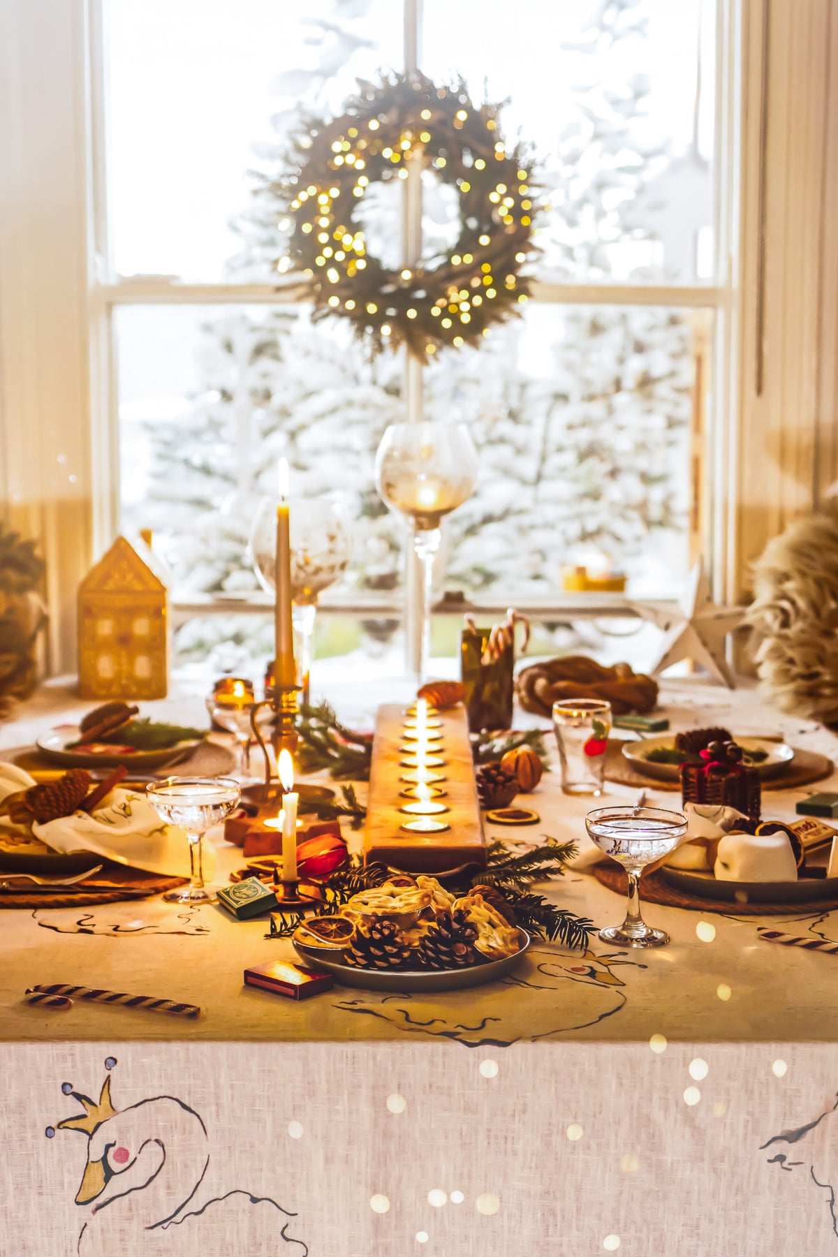 A beautifully decorated Christmas dining table set with candles, plates of food, wine glasses, pinecones, and holiday greenery, with a glowing wreath hanging in the background near a bright window.