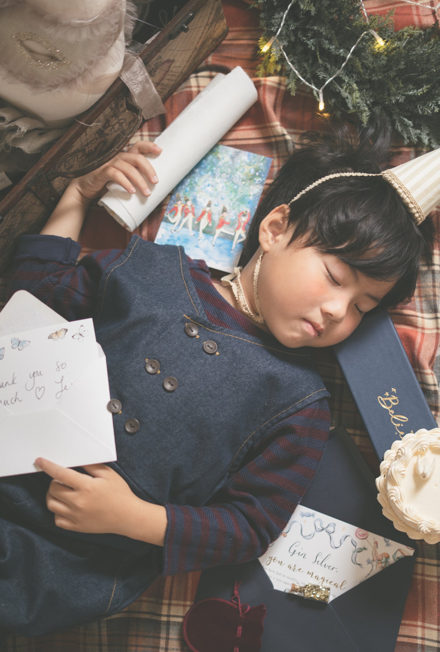 A young child wearing a navy blue denim vest over a red striped shirt lies asleep on a red plaid blanket, surrounded by Christmas-themed items including a gift box, card, and evergreen branches.