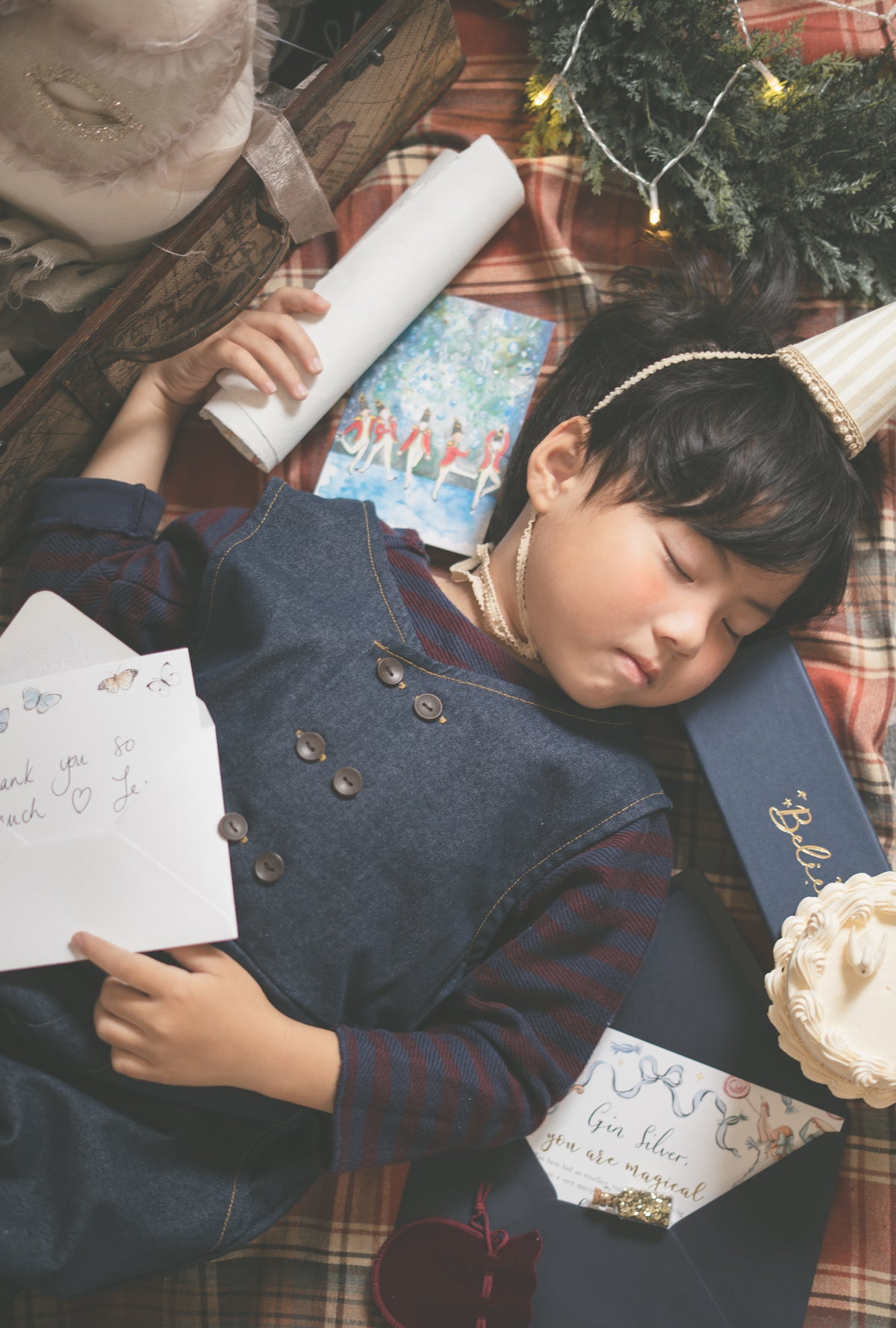 A young child wearing a navy blue denim vest over a red striped shirt lies asleep on a red plaid blanket, surrounded by Christmas-themed items including a gift box, card, and evergreen branches.
