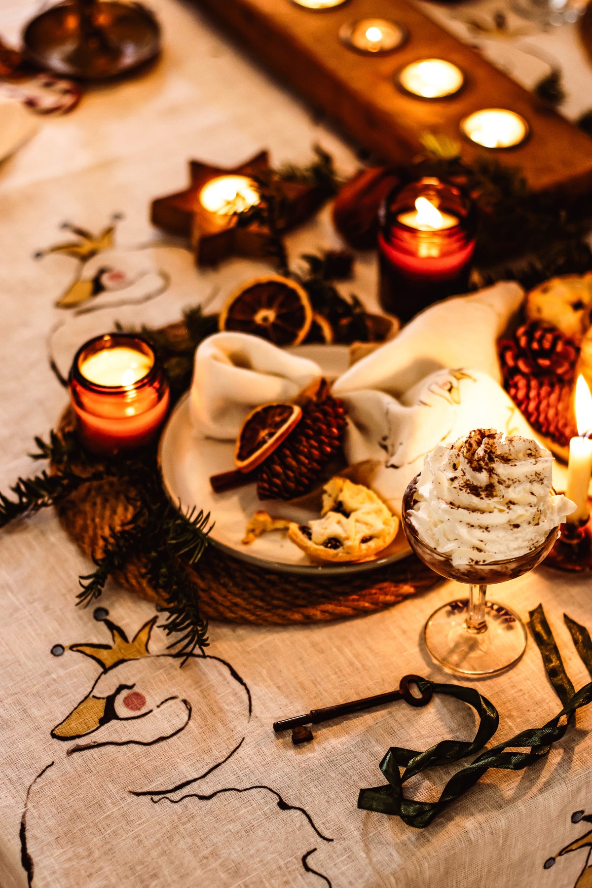 A cozy tabletop scene with a dessert in a stemmed glass, cookies on a plate, candles, pinecones, dried orange slices, and embroidered table linens under warm candlelight.
