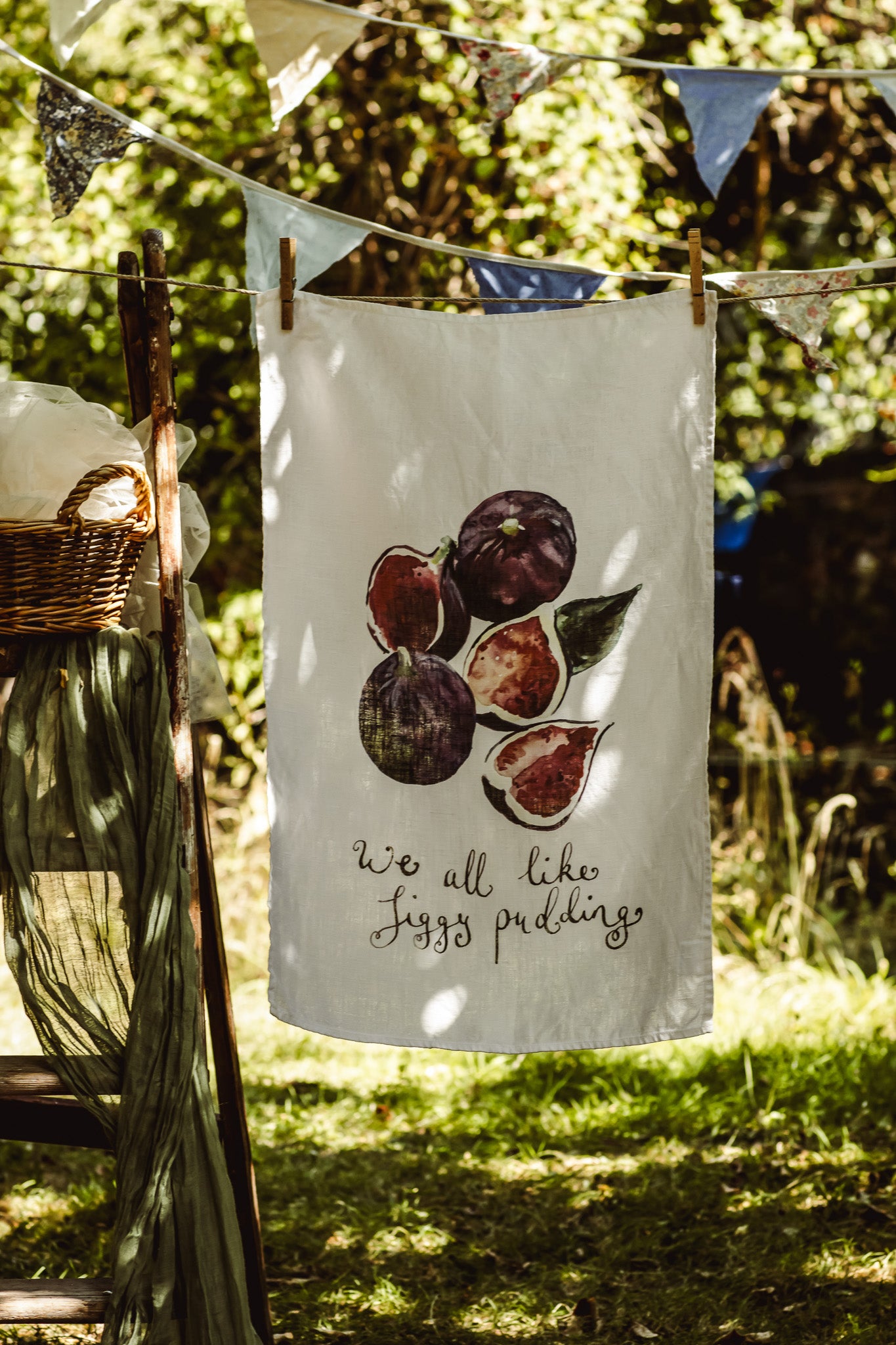 A white kitchen tea towel with a watercolor print of figs and the text 'we all like figgy puddings' hanging from a clothesline outdoors in a garden setting.