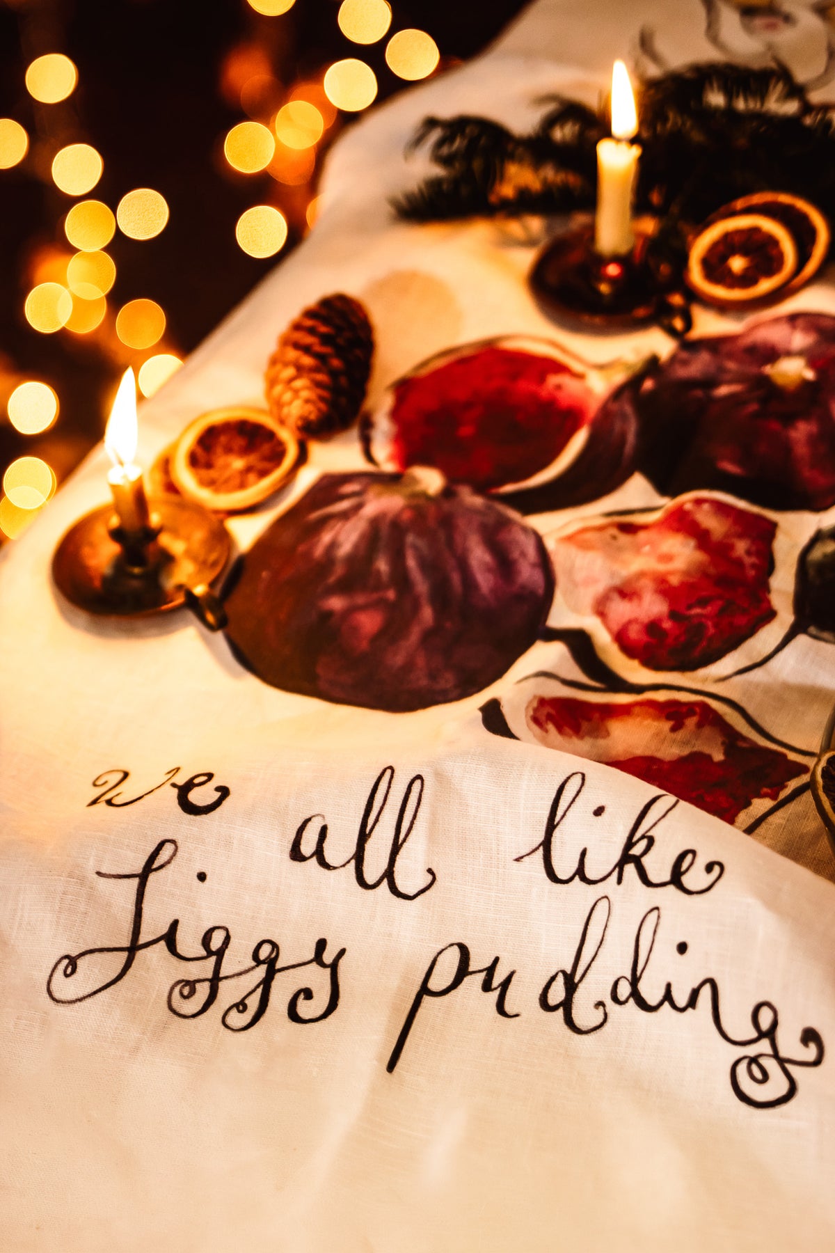 A close-up of a white fabric napkin or table runner with elegant black cursive script reading 'all like Jiggy pudding' and printed illustrations of figs and dried orange slices, styled with candles and warm bokeh lights.