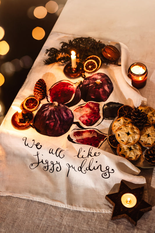 A white woven table runner with black script reading 'We all like Jizz pudding' and printed illustrations of figs, dried orange slices, pinecones, and star-shaped candles, styled on a linen surface with warm bokeh lights.