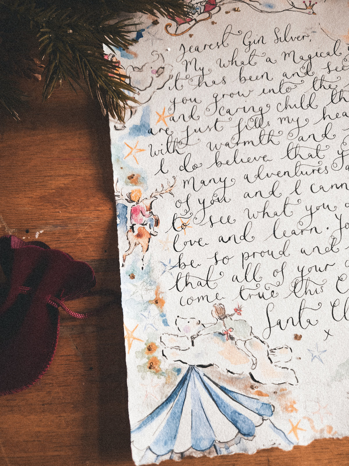Hand-lettered Christmas card on textured handmade paper, styled on a wooden surface with pine needles and a burgundy bow.