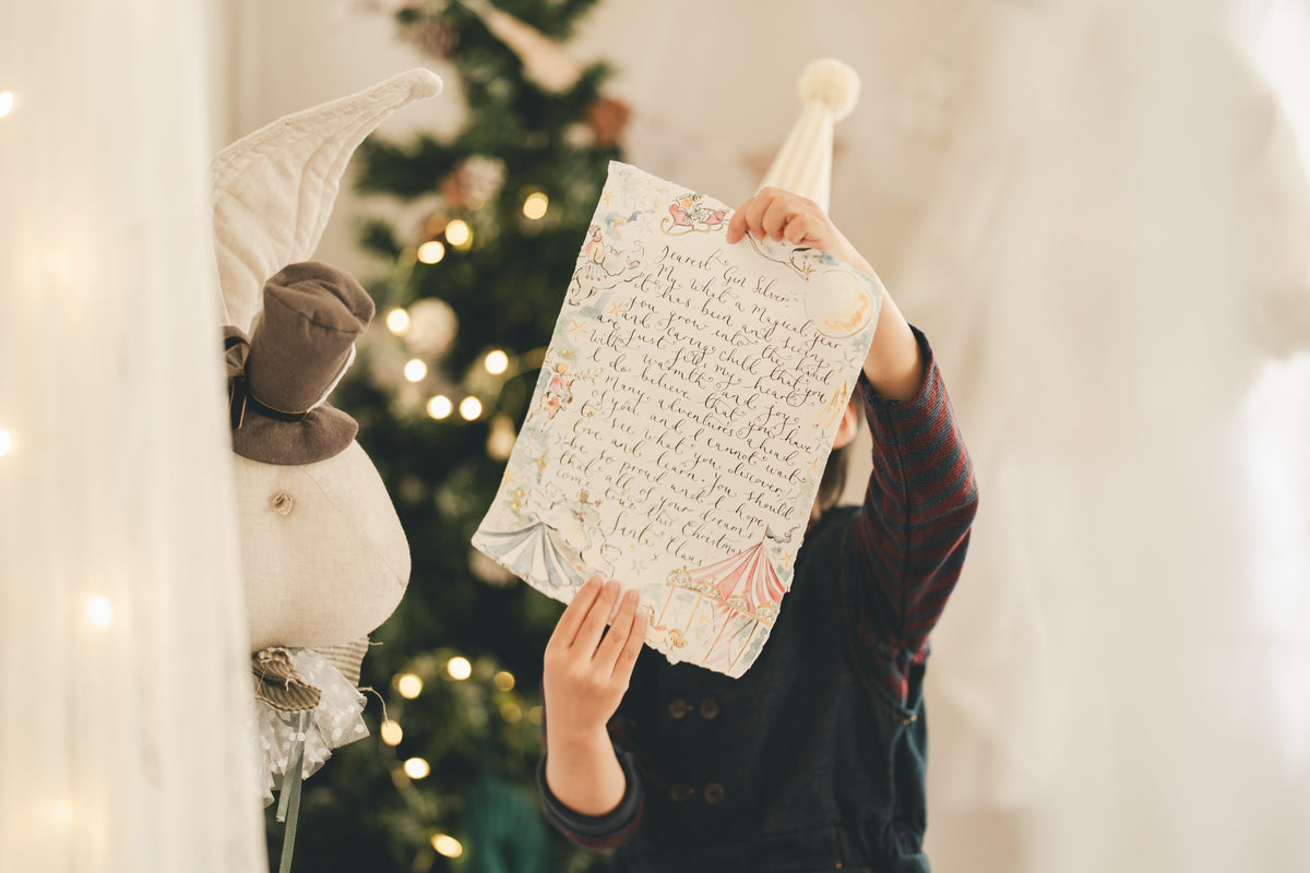 A child holds up a decorative letter card with cursive handwriting and pastel illustrations, posed in front of a softly blurred Christmas tree with warm lights. A plush holiday decoration is visible on the left.